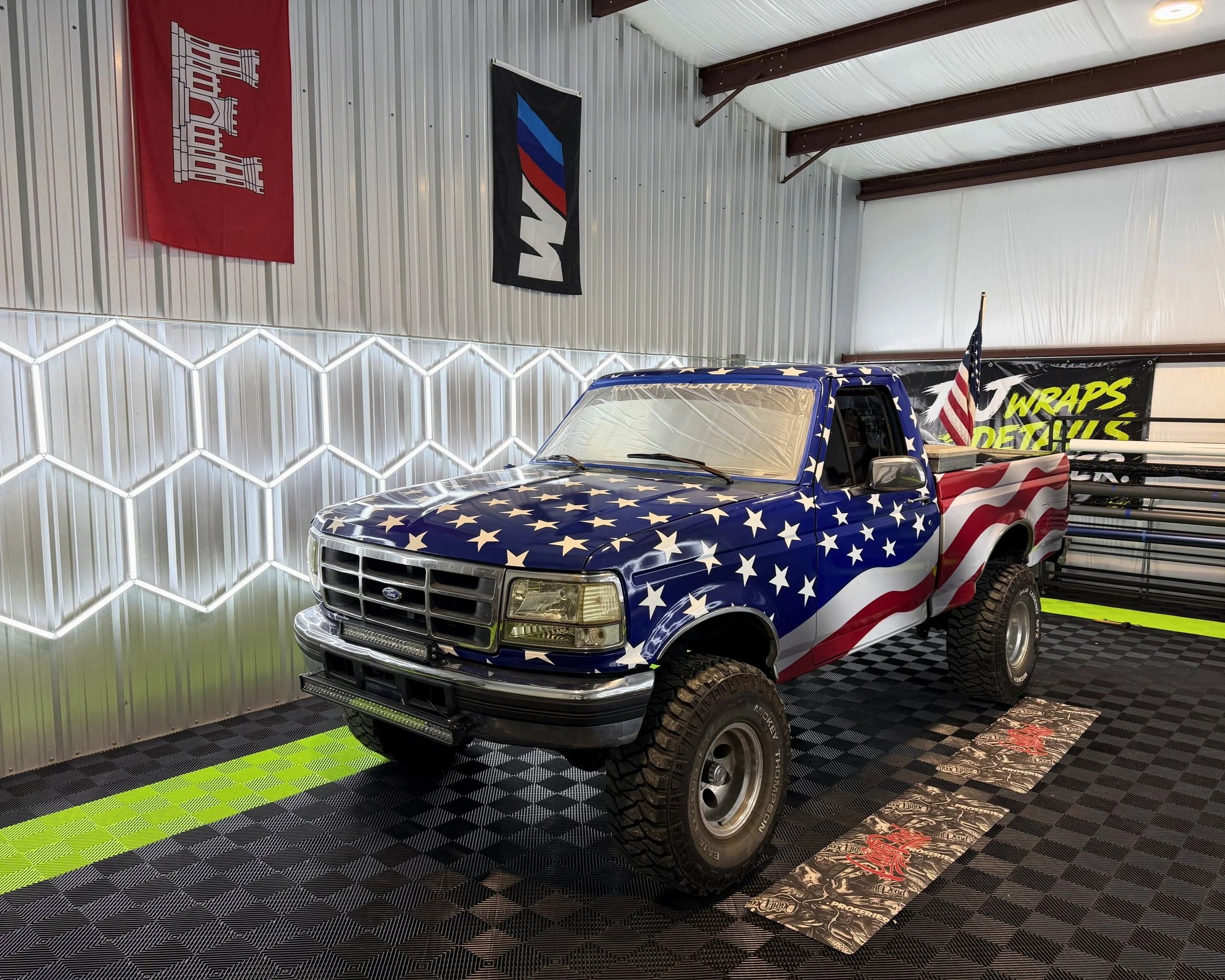 A pickup truck with an American flag theme paint job inside a metal-walled garage with flags on the wall and bright lighting.