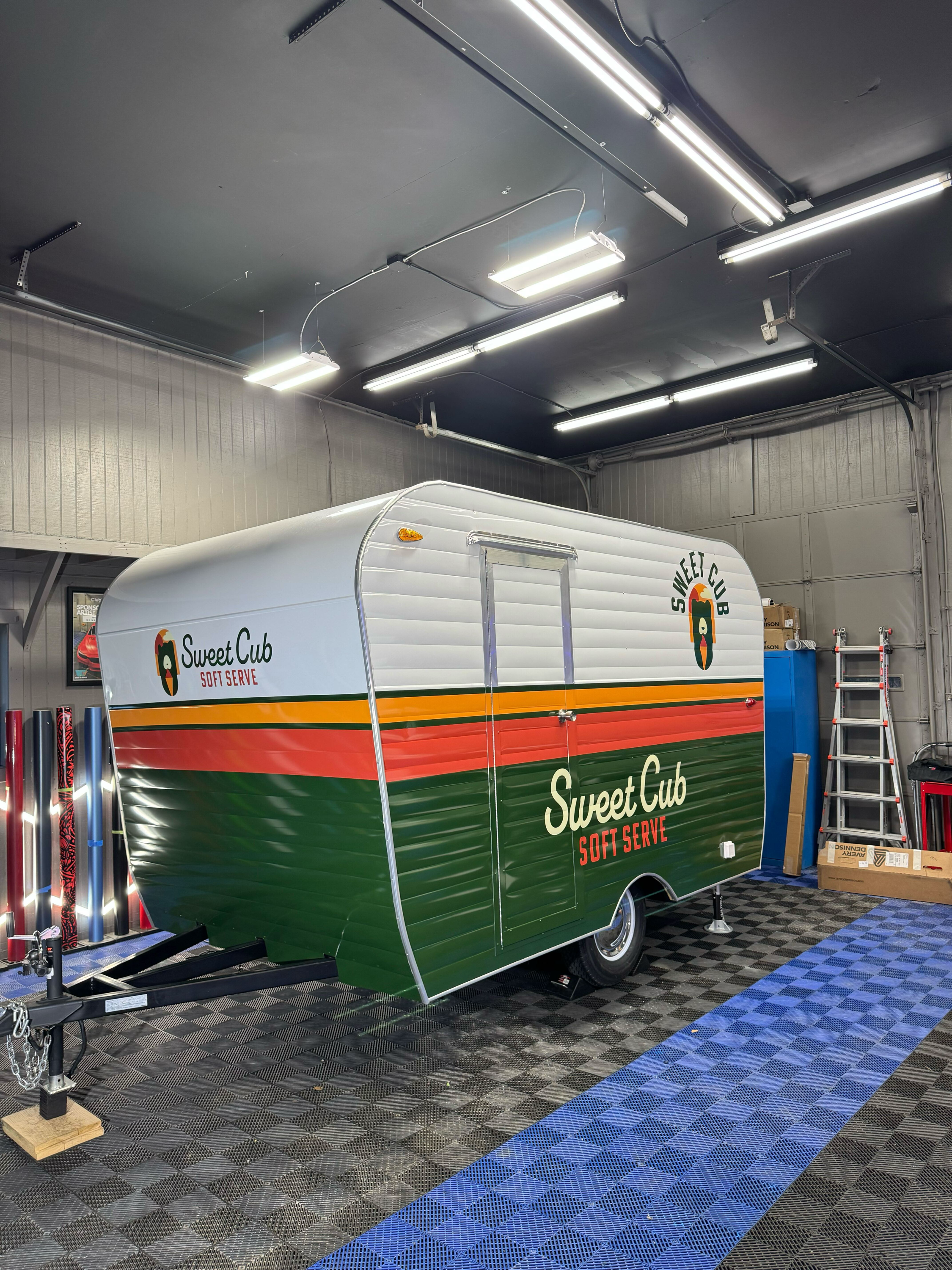 A white food trailer with green, orange, and red stripes and the logo 'Sweet Cub' and the words 'Soft Serve.' It is inside a garage or workshop with black and gray patterned flooring, illuminated by fluorescent ceiling lights, with tools and equipment nearby.