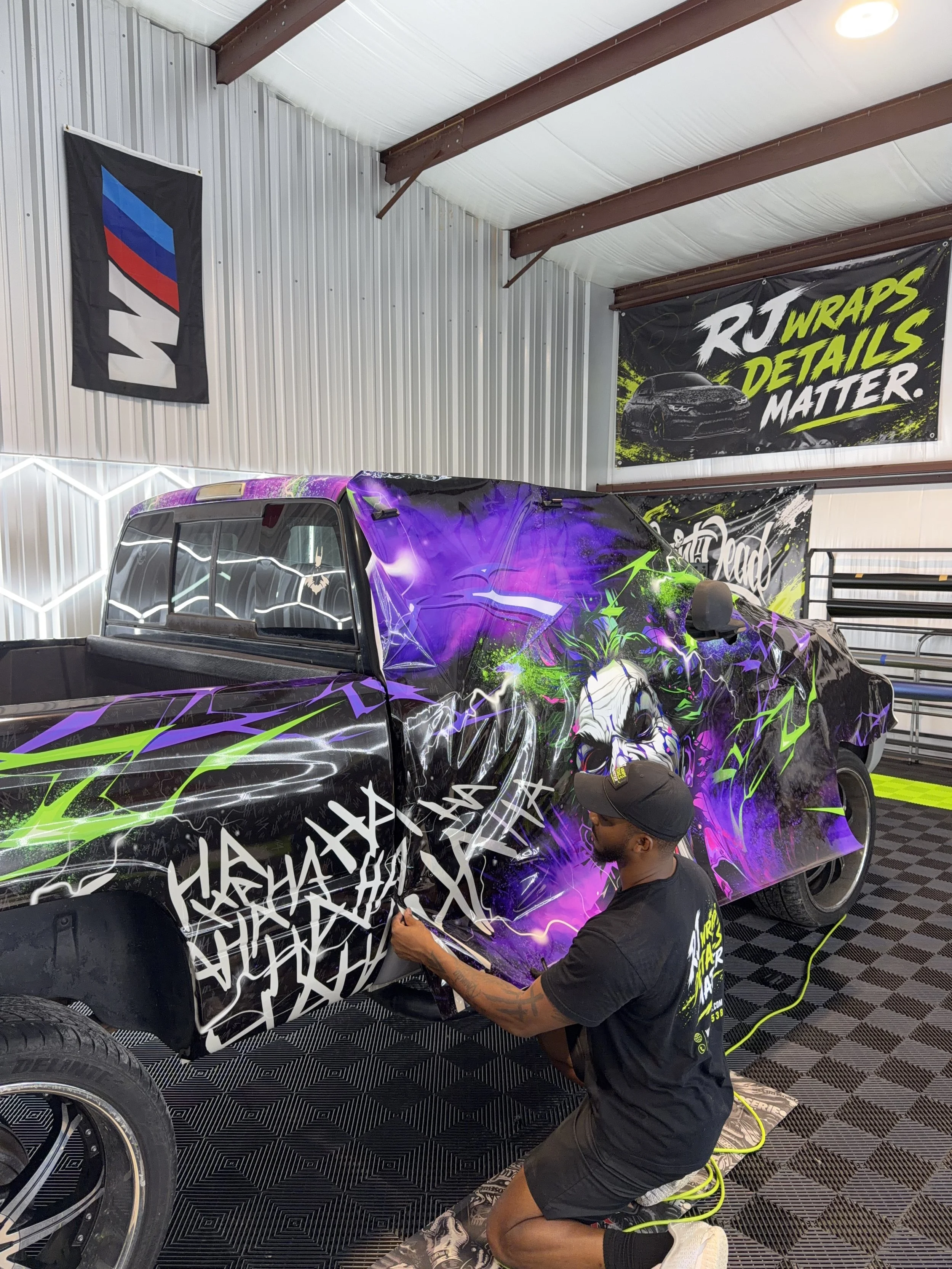 A man is wrapping a black pickup truck with a vibrant custom vinyl wrap featuring purple, green, and white graphic designs, in a garage with banners on the wall that say 'RJ Wraps' and 'Details Matter'.