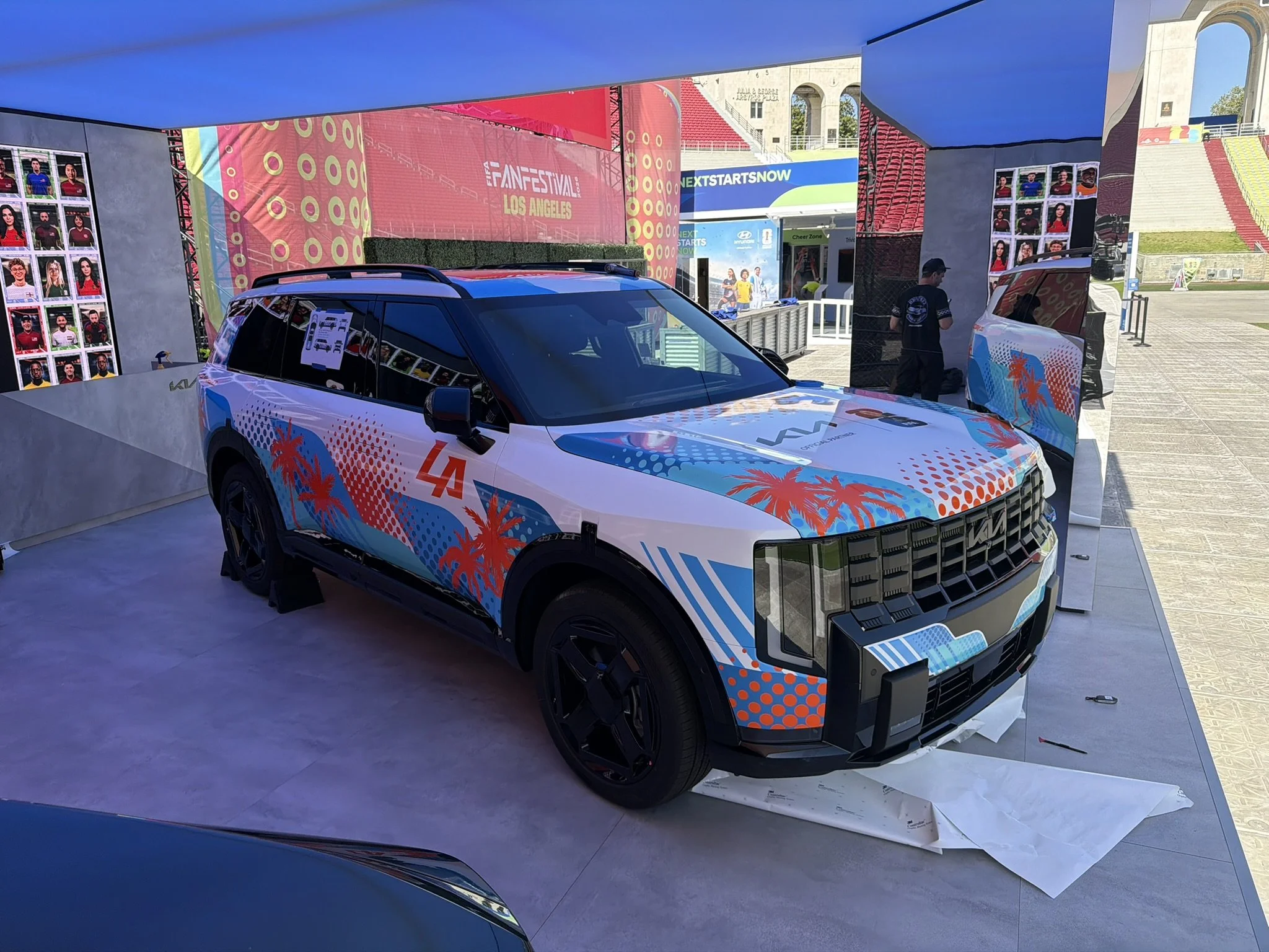 An outdoor event setup with a white SUV featuring colorful graphics of palm trees and a sunset, positioned under a large blue tent. Behind the vehicle are display walls with photos of soccer players, and banners promoting the FIFA Fan Festival in Los Angeles.