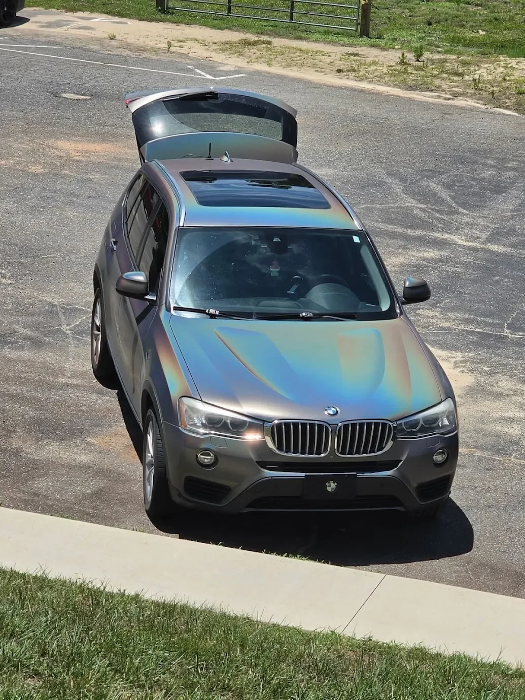 A silver BMW SUV with a rainbow-colored reflective surface parked on a gravel lot, with its rear hatch open and a grassy area in the foreground.
