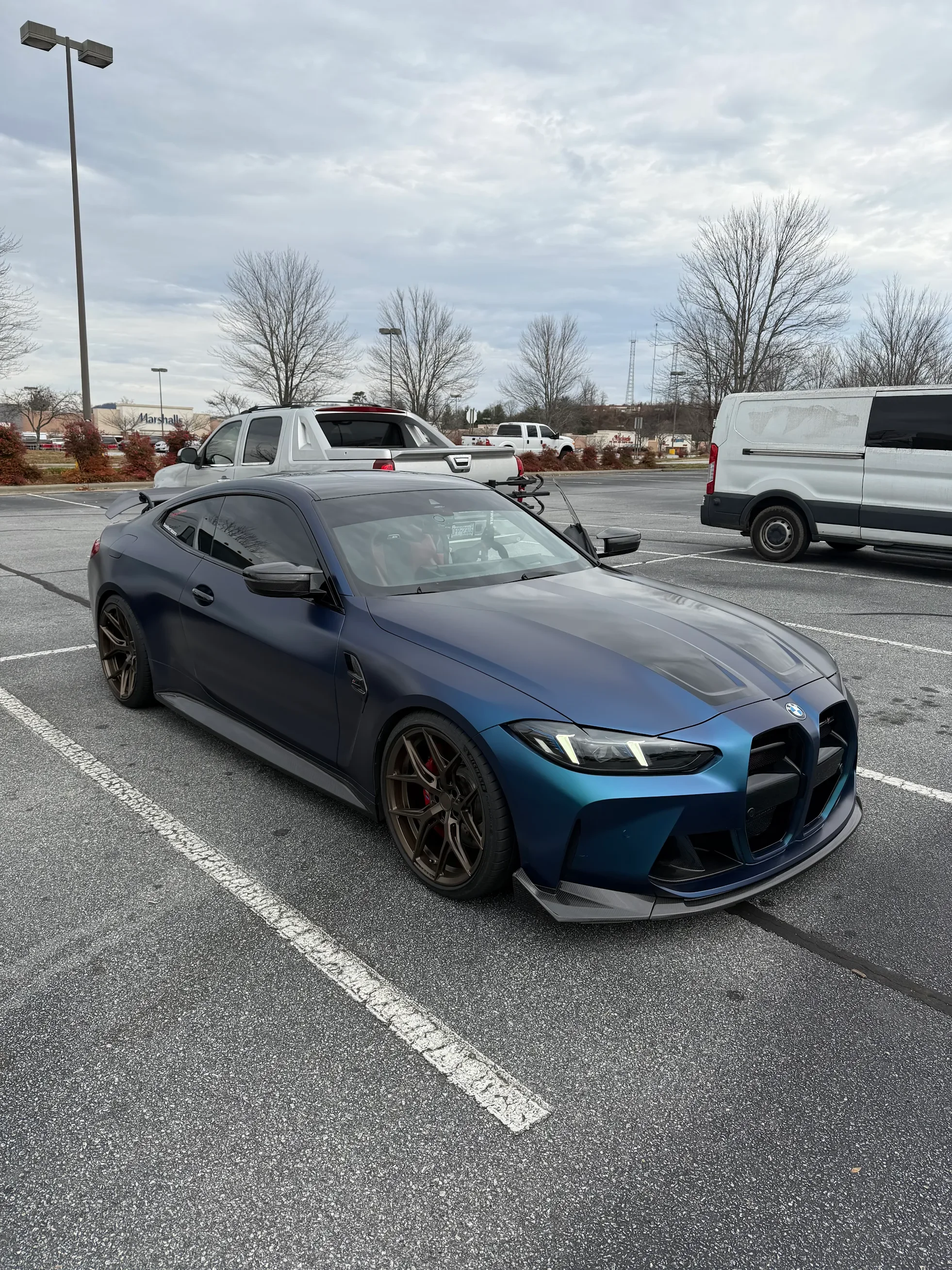 A dark blue BMW sports car with black wheels parked in a parking lot, with a silver pickup truck and a white cargo van nearby, and leafless trees in the background under a cloudy sky.