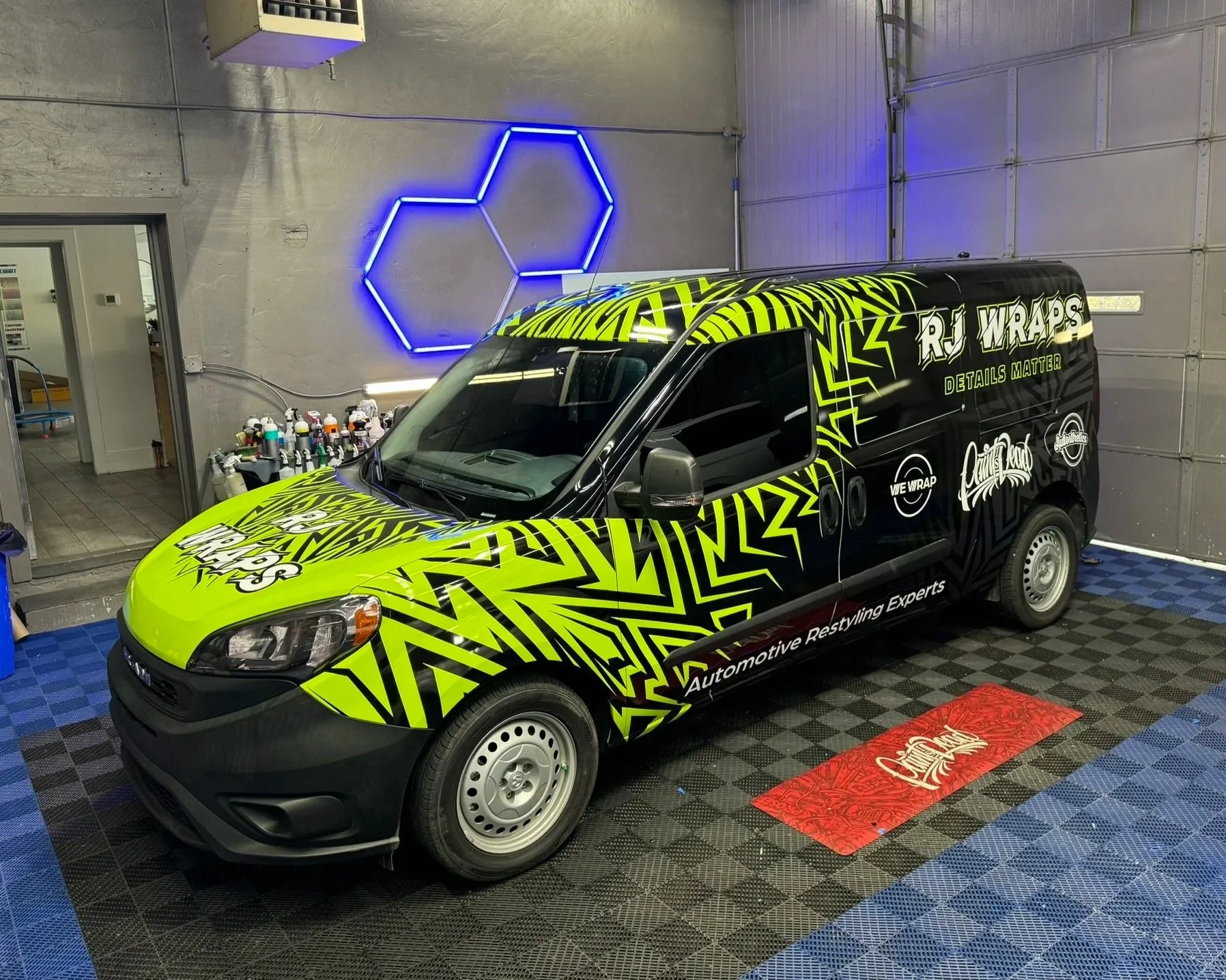 A black van decorated with bright yellow and green graphics and the words "RJ WRAPS" and "Automotive Restyling Experts" parked inside a garage. The garage has a geometric neon light on the wall, a shelf with spray paint cans, and a partially open door leading to an adjacent room. The floor has a black and blue checkered pattern with a red mat in front of the van.