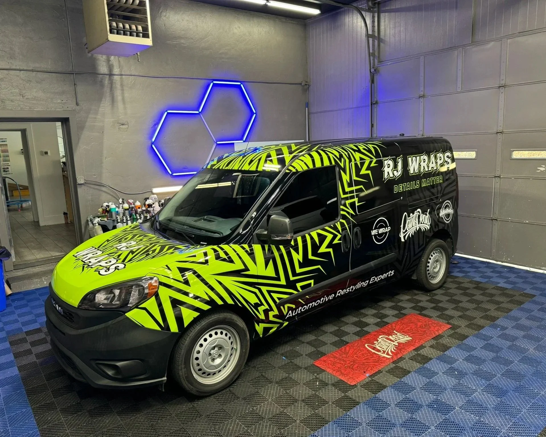 A black van with green and yellow graphic design, parked inside a garage. The van has text that reads 'RJ Wraps' and 'Automotive Restyling Experts.' Behind the van, a wall features a blue neon hexagon design, and there are shelves with spray paint bottles and a workstation nearby.