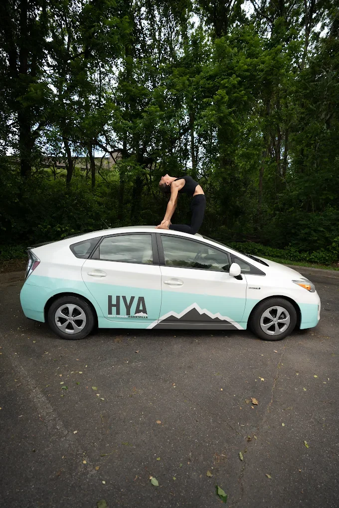 A woman is doing yoga on the roof of a white and light blue electric car with Mountain Asheville logo and HYA text, parked outdoors near trees.