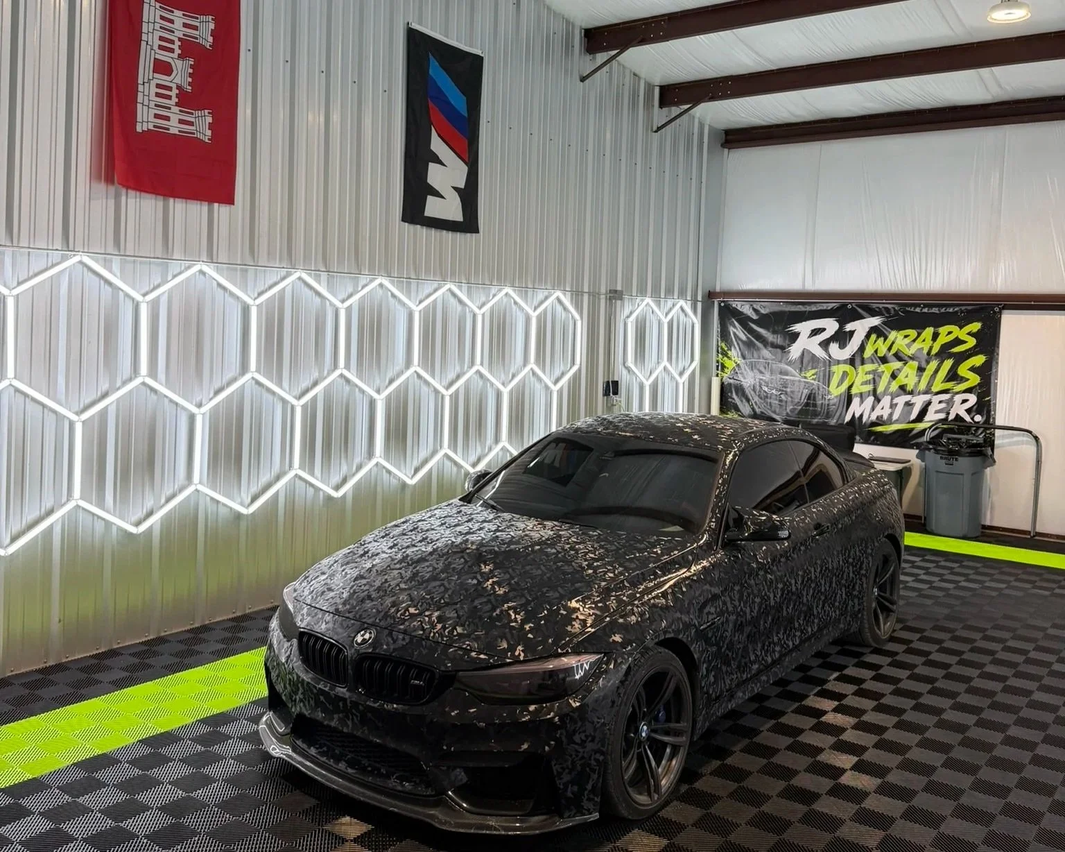 Black sports car with camouflage wrap parked inside a detailing shop with banners on the wall reading 'RJ Wraps' and 'Details Matter', neon light hexagon pattern on the wall, checkered floor, trash can and equipment.