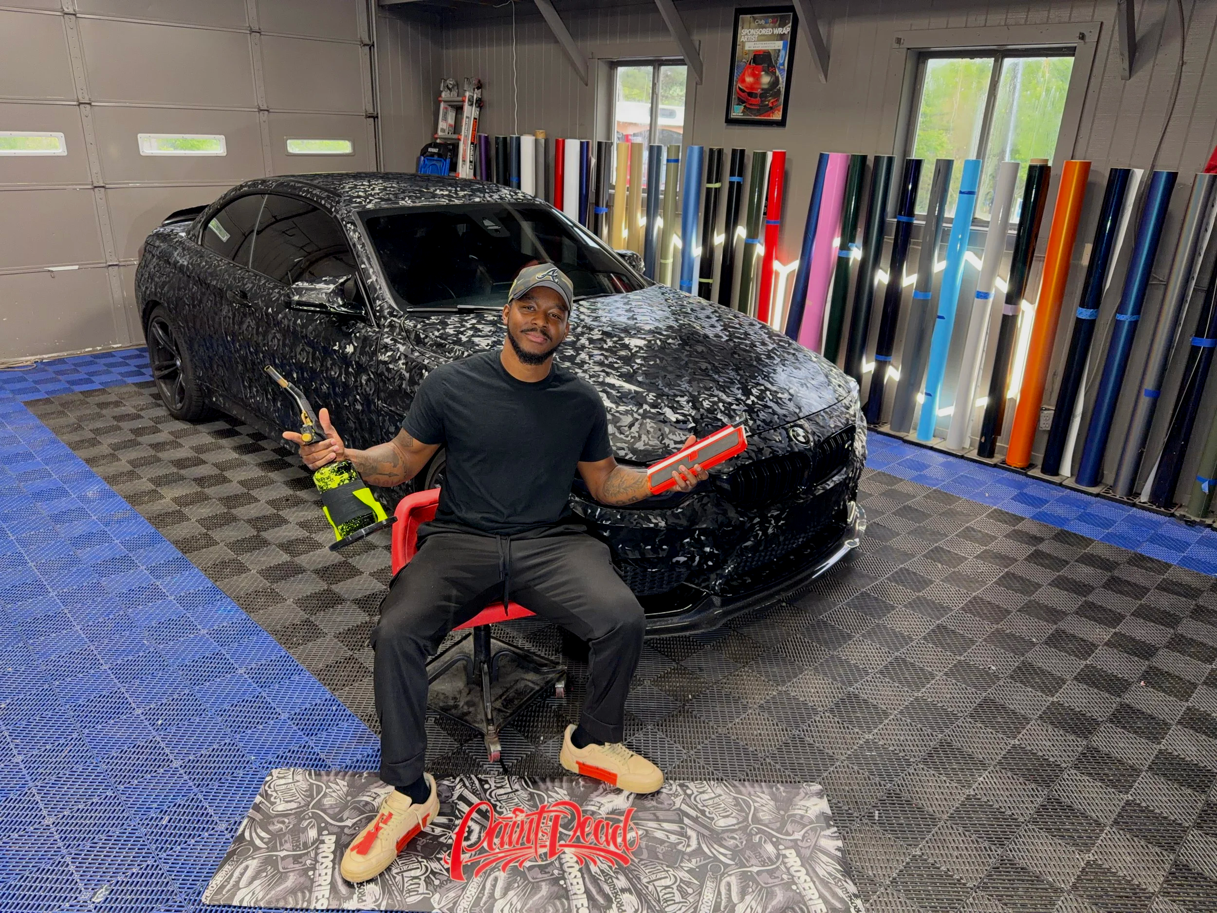 A man sitting inside a garage with a black sports car covered in camouflage wrap behind him. He is holding a drill and a red tool box, wearing a black t-shirt, black jeans, and beige sneakers. There are colorful wraps or decals stored vertically in the background and a 'Saints Dead' branded mat on the floor. The garage has a gray and blue checkered floor and windows on the walls.