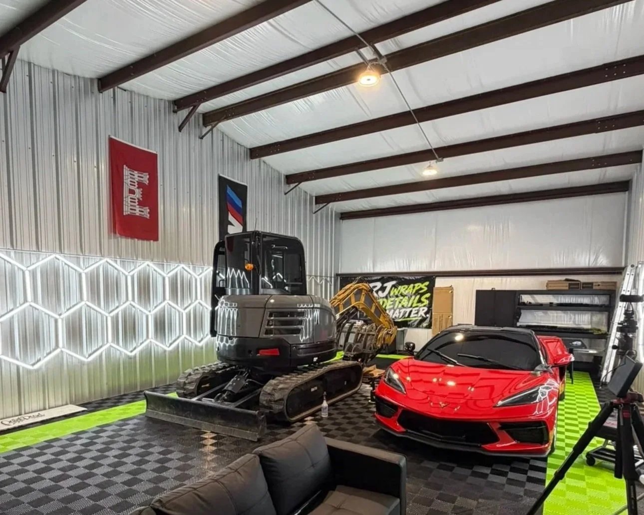 An indoor garage or workshop space with a black tracked mini excavator and a red sports car parked inside. The room has metal walls, a high ceiling with exposed beams, and decorative lighting. There are banners on the wall and a black storage shelf in the background.