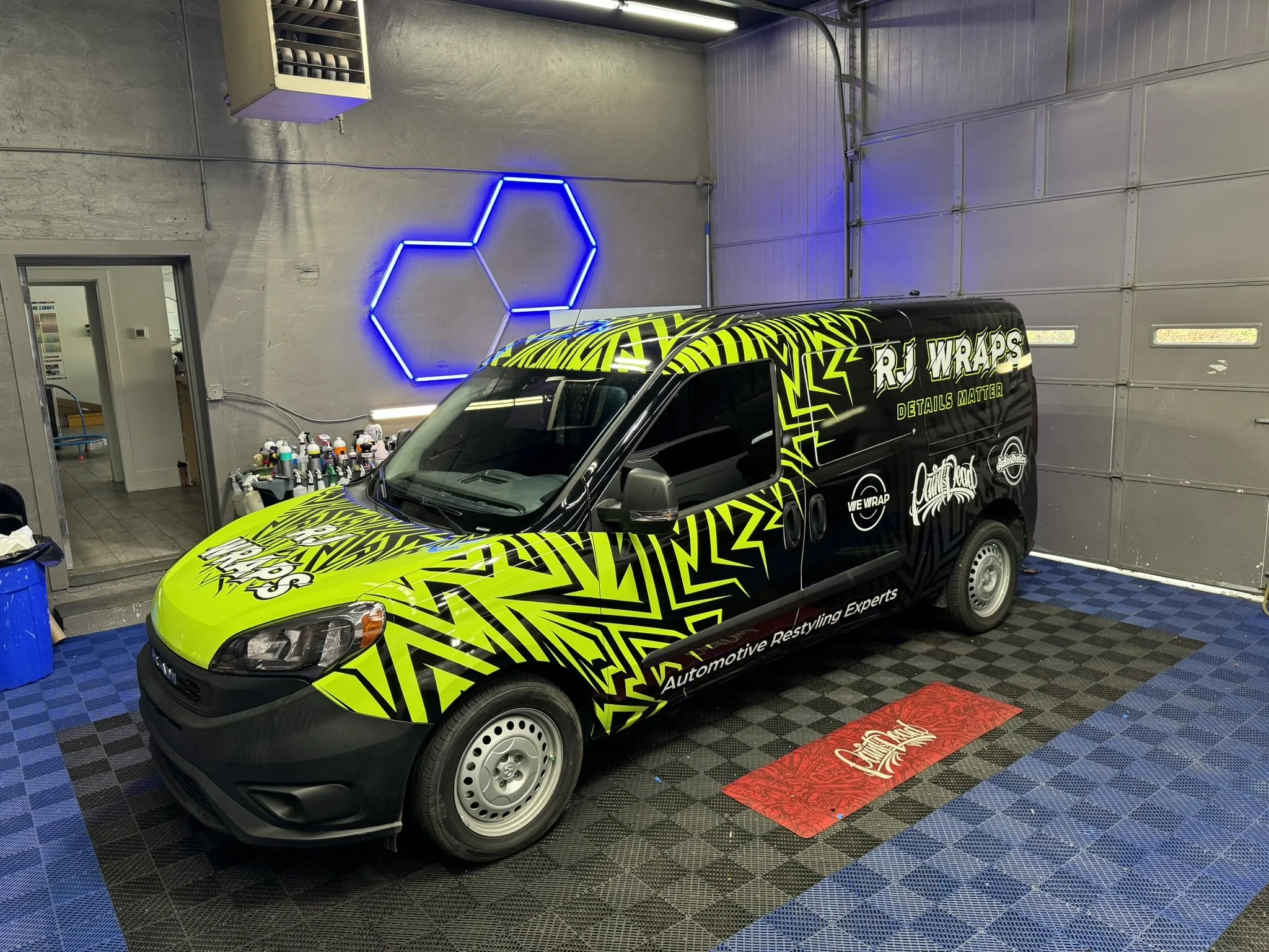 A black van with bright yellow and black graphics and lettering, parked inside a garage. The van has the logos 'RJ Wraps' and 'We Wrap' on its side. The garage interior features a neon blue geometric light on the wall, a work table with spray paint cans, and a textured blue floor with a red mat near the entrance.