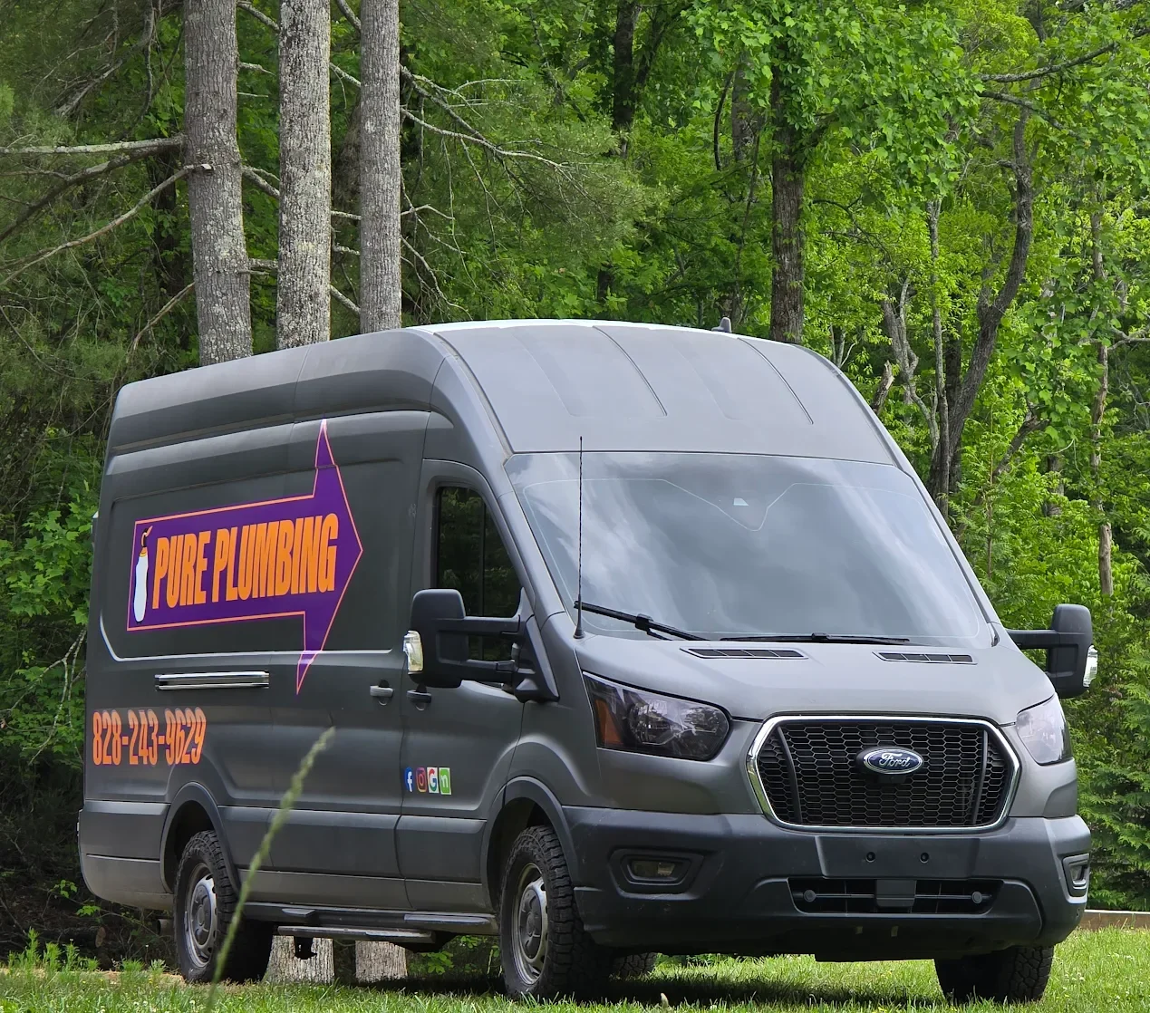 Gray Ford transit van with signs for Pure Plumbing parked on grass with green trees in the background.