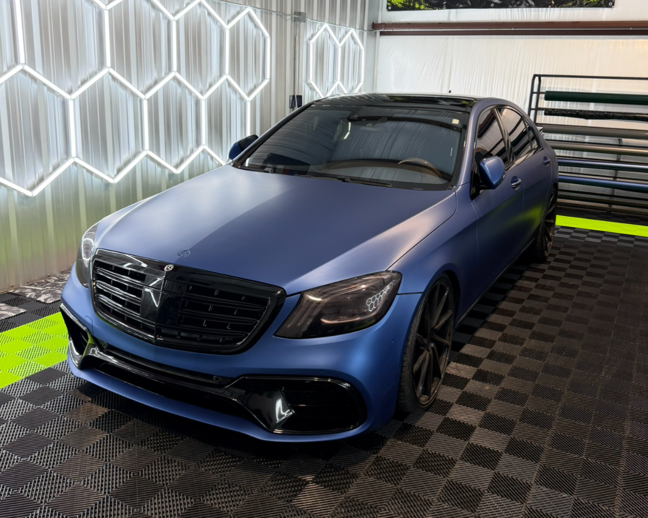 Blue luxury sedan with black grille and black wheels inside a garage with geometric wall lighting and checkered flooring.