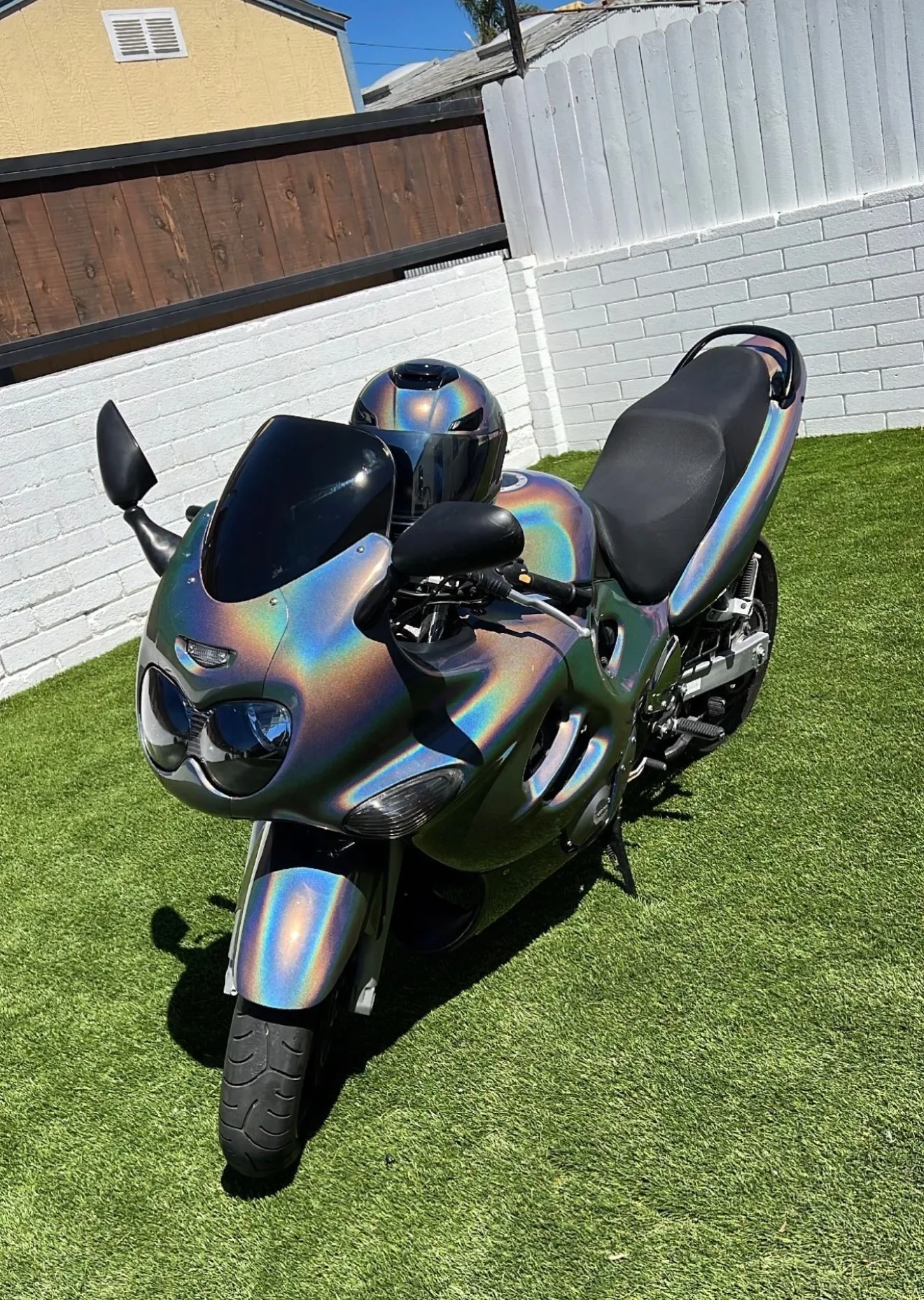 A motorcycle with a rainbow-colored, holographic paint job parked on a lawn, with a white brick wall and a yellow building in the background.
