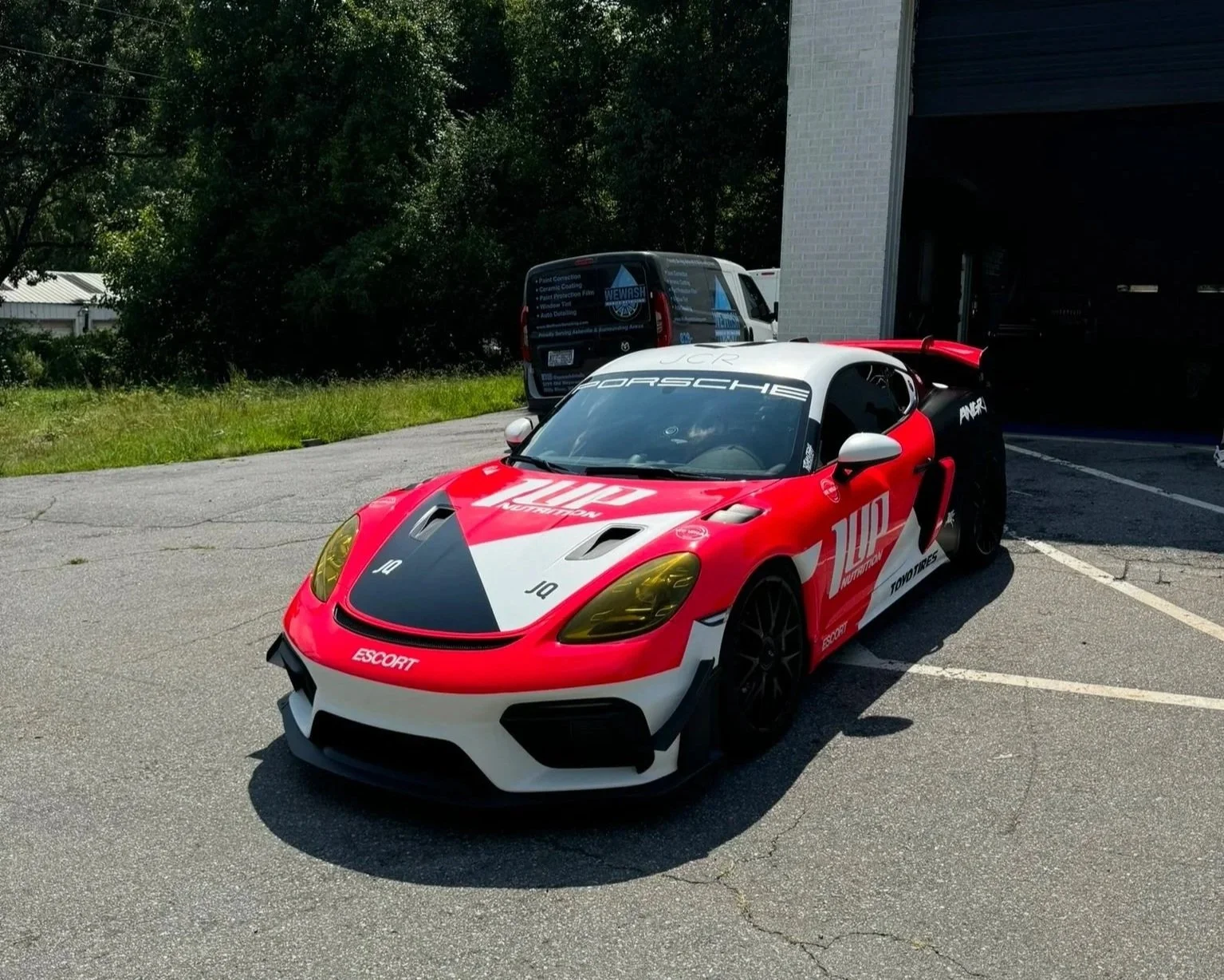 A red, white, and black Porsche race car parked in a parking lot with some trees and a building in the background.