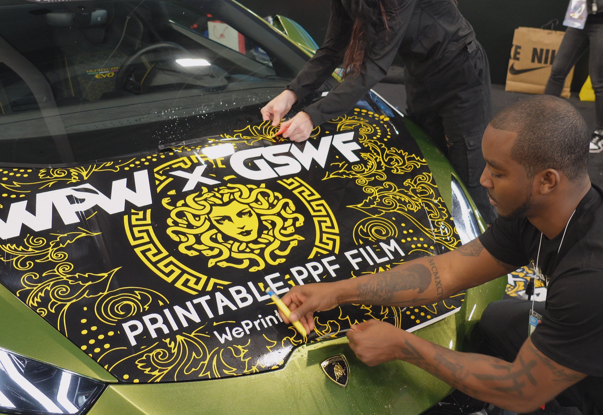 Two people working on a vehicle wrap, applying a black and yellow vinyl design with the words 'PRINTABLE: WePrint' on the hood of a green Lamborghini.
