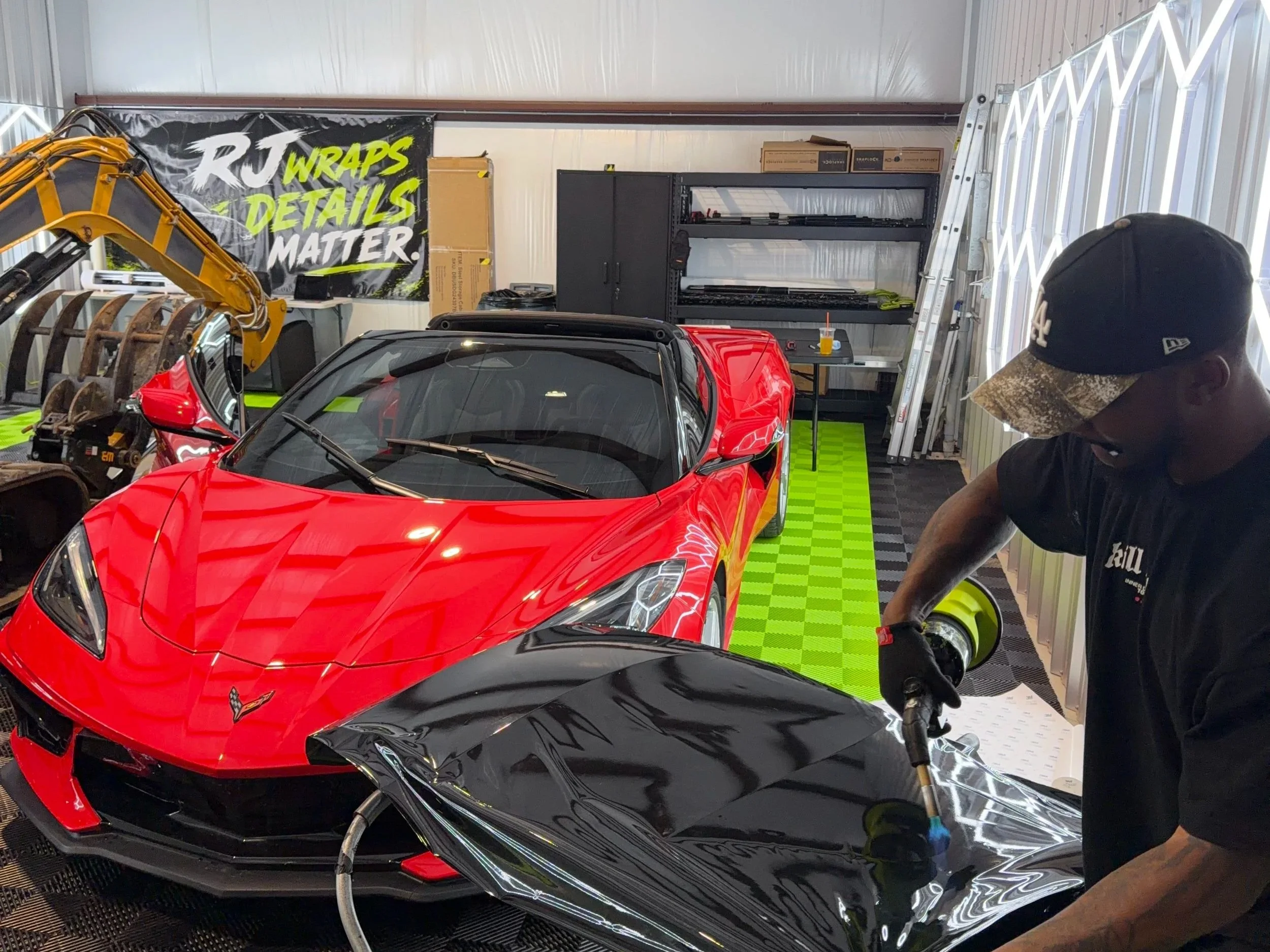 A red sports car parked inside a garage with black and green checkered floor. There is a man in a camouflage cap working on the black part of the car. Garage shelves, ladder, tools, and a large banner that reads 'RJ WRAPS DETAILS MATTER' are visible in the background.