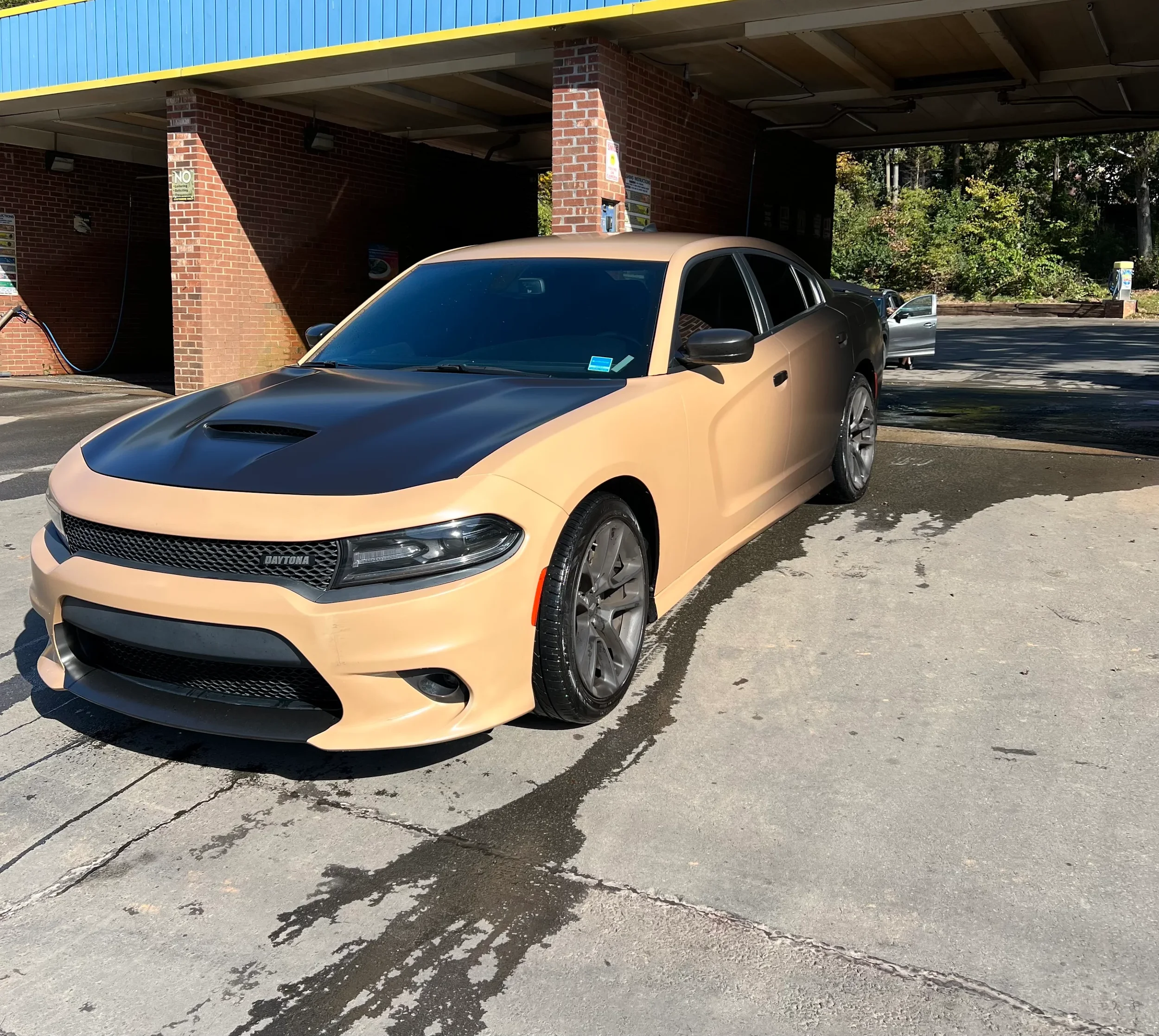 A beige and black sports car with a Daytona logo on the front, parked at a car wash station with red brick columns and a garage overhang, surrounded by trees and a parking lot.