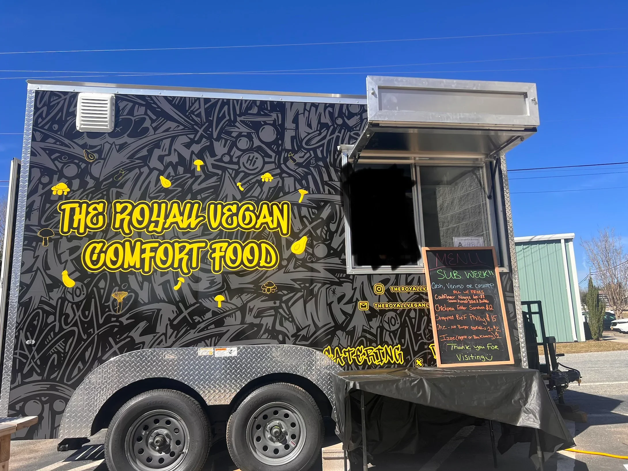 Food truck with black graffiti art design, yellow lettering that reads 'The Royal Vegan Comfort Food,' and a blackboard menu listing various vegan items.