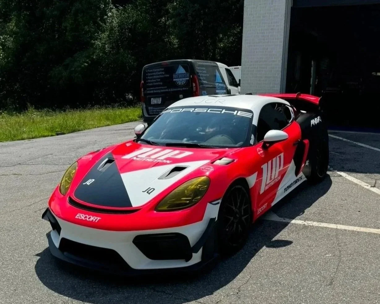 A red, white, and black race car with Porsche branding parked in a parking lot. The car has yellow-tinted headlights and various sponsor logos, including 100 Throttle and Toyo Tires.