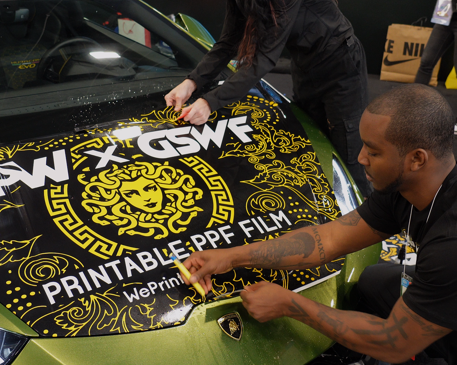 Two people applying a vinyl decal with black and yellow design containing a Medusa head logo and text onto the hood of a green Lamborghini sports car.