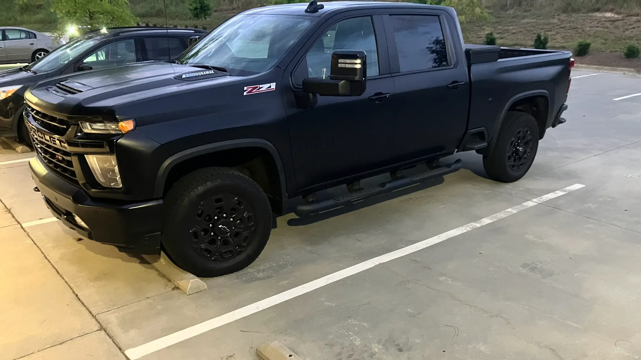 A black Chevrolet Silverado pickup truck parked in a parking lot at dusk with grass and trees in the background.