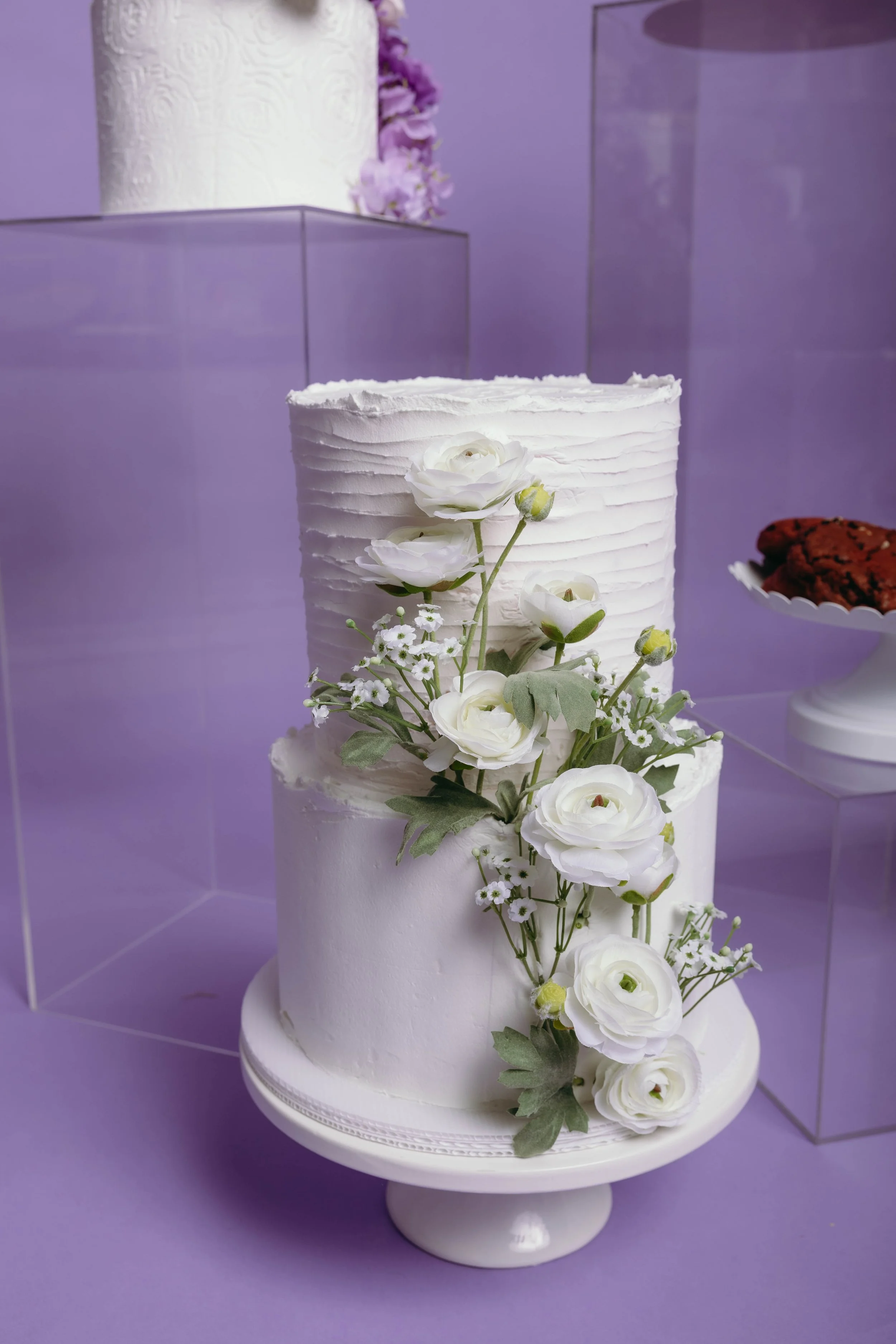 A three-tier white wedding cake decorated with white roses, lisianthus, and greenery, placed on a white cake stand against a purple background.