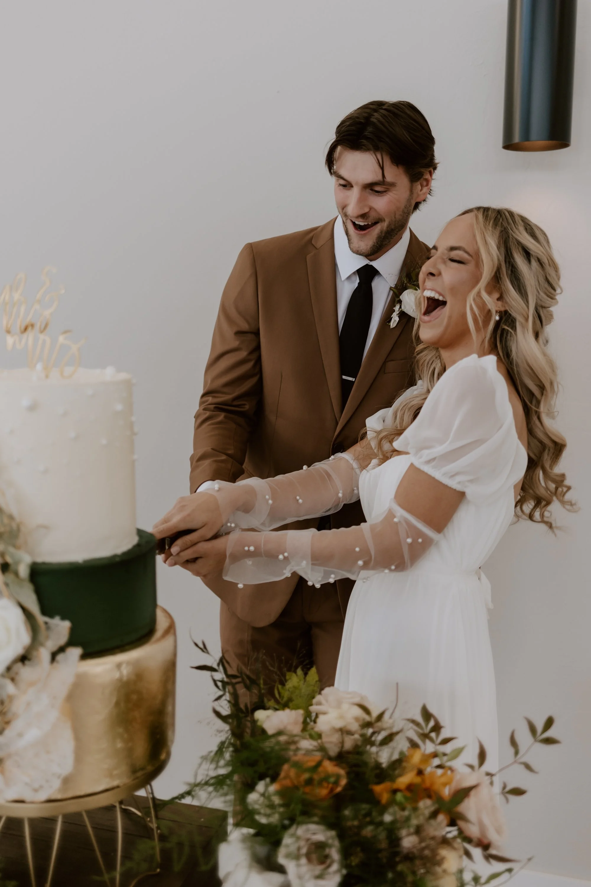 A man and woman in wedding attire celebrating as they cut a wedding cake together at their wedding reception. The man is wearing a brown suit and the woman is wearing a white dress with sheer, pearl-embellished sleeves.