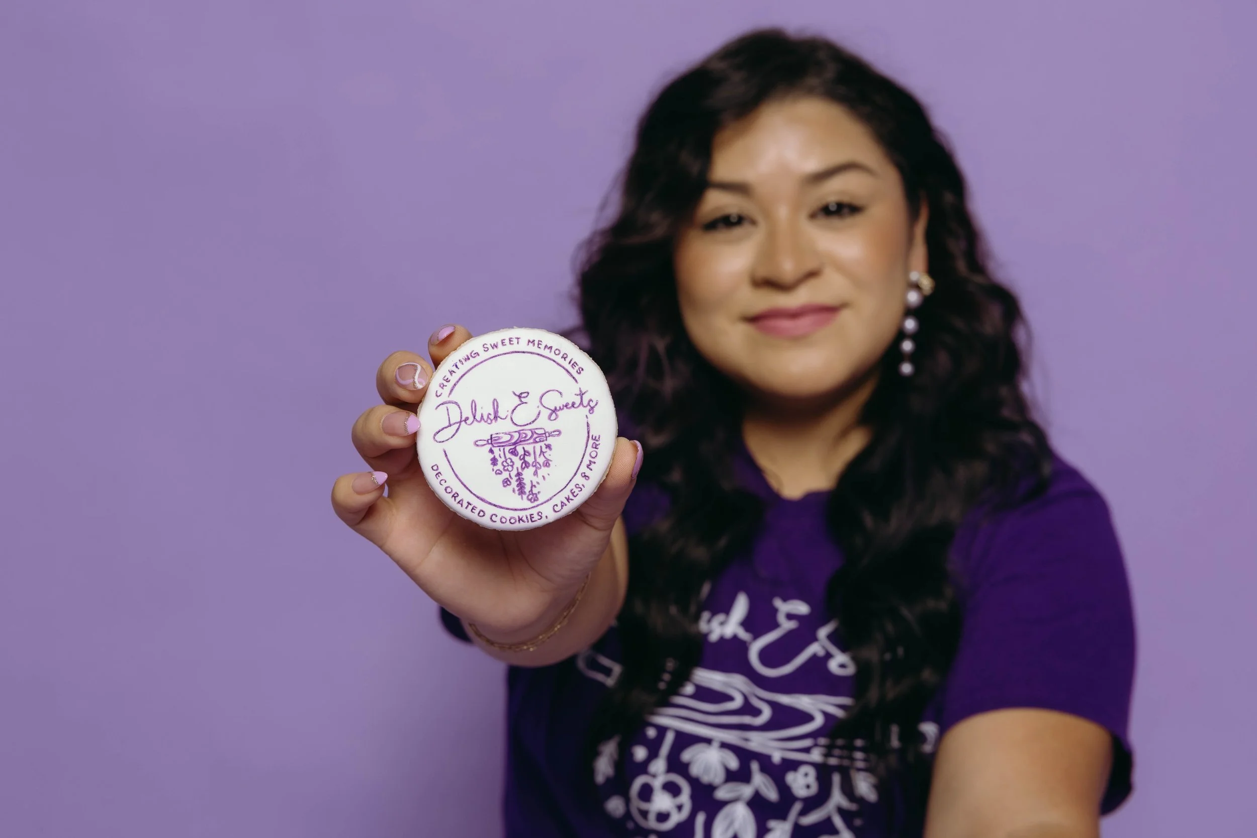 The Owner Anicia Vasquez with dark, wavy hair and pearl earrings holds a round cookie toward the camera. The cookie has purple writing that says "Delish E Sweets." The background is purple, and she is wearing a purple shirt with white lettering.