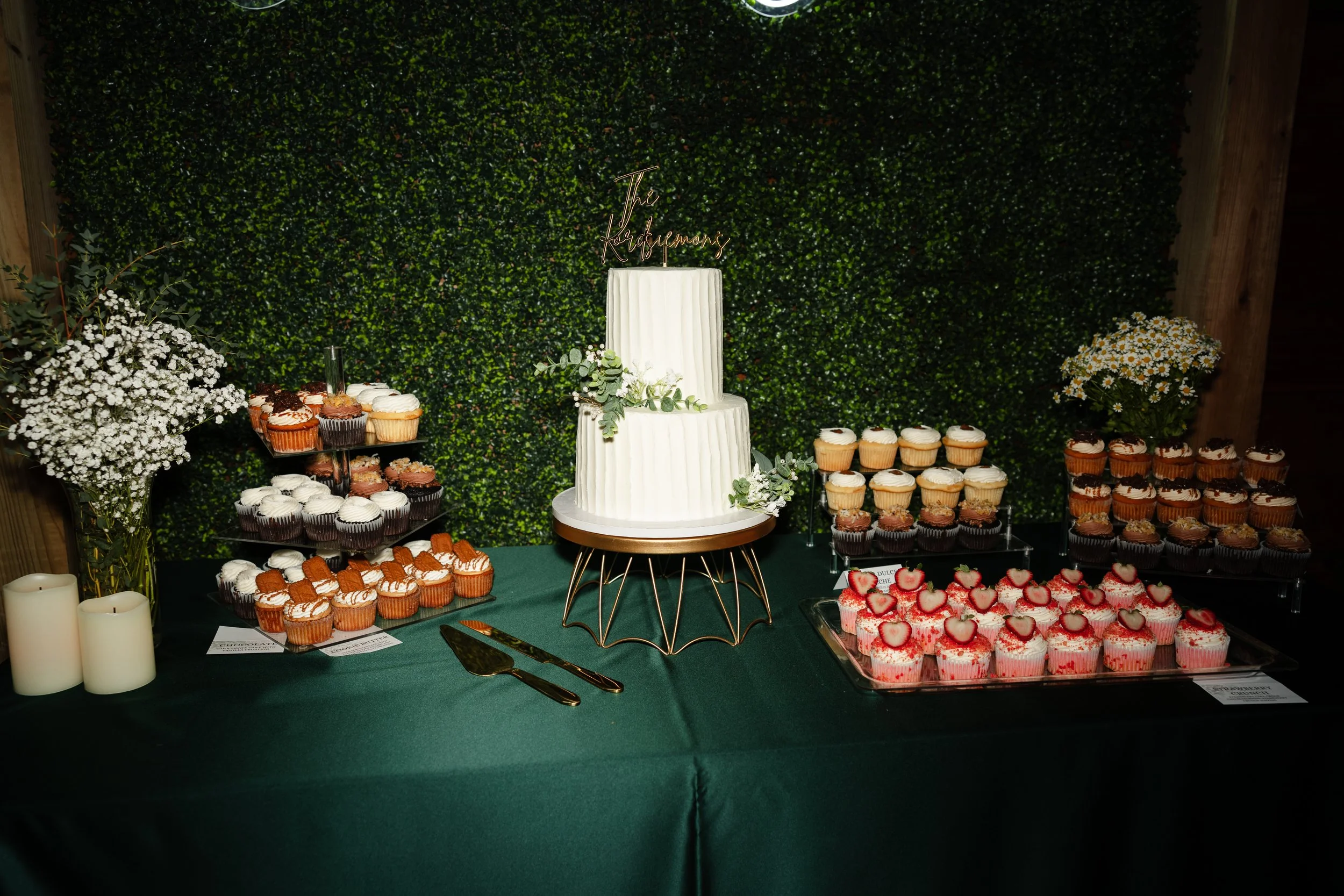 Decorative table with an assortment of cupcakes, a white-tiered cake with greenery and a gold cake topper reading "The Kershyers," and floral arrangements on a green tablecloth against a green leafy wall backdrop.