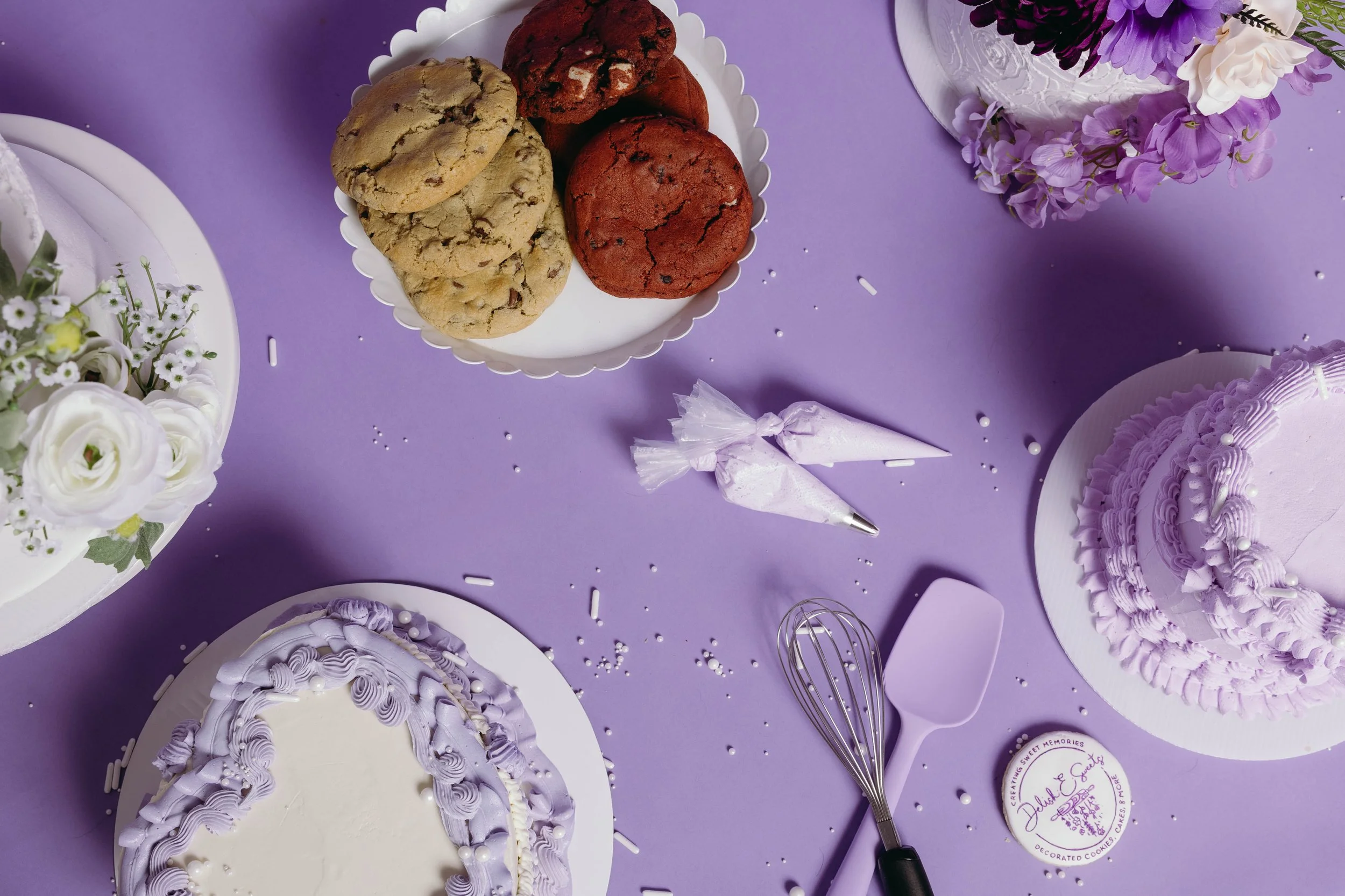 A white plate with assorted cookies, purple decorated cakes, and flower arrangements on a purple table. There are also cake decor tools and confetti scattered around.