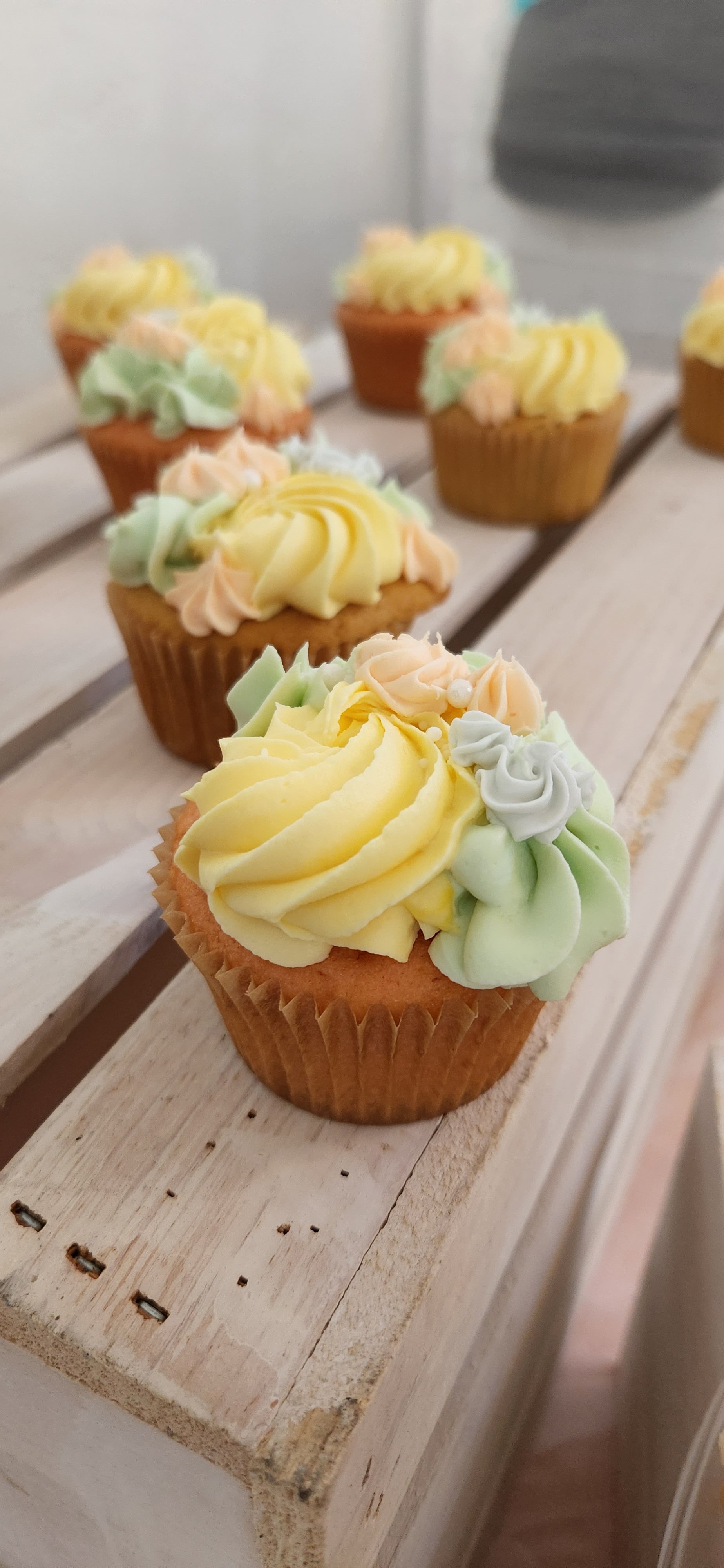 Decorated cupcakes with pastel yellow, green, pink, and white frosting on a white wooden surface.