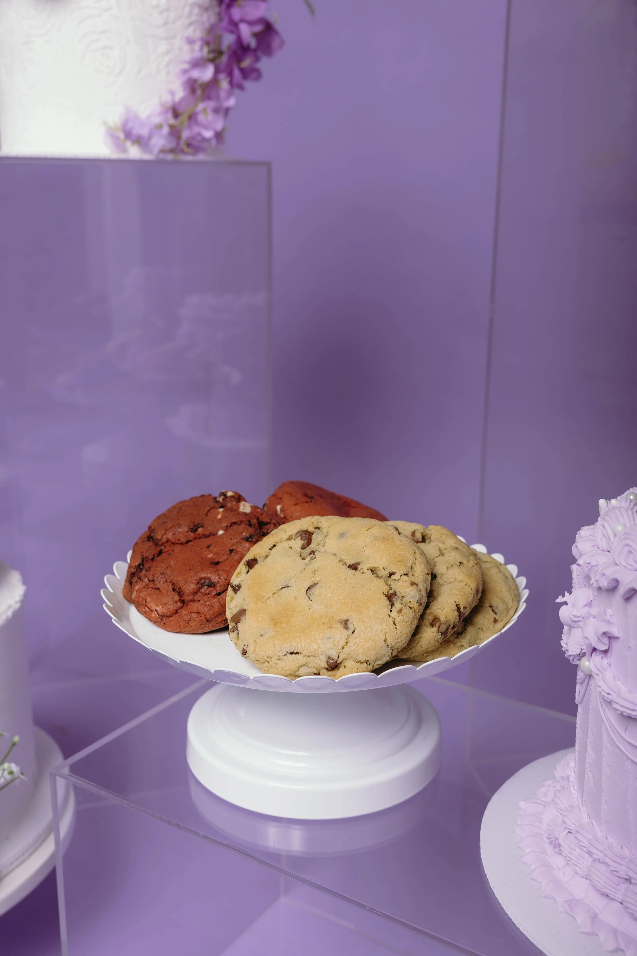 A white cake stand with chocolate chip cookies on top, displayed against a purple background with purple flowers.