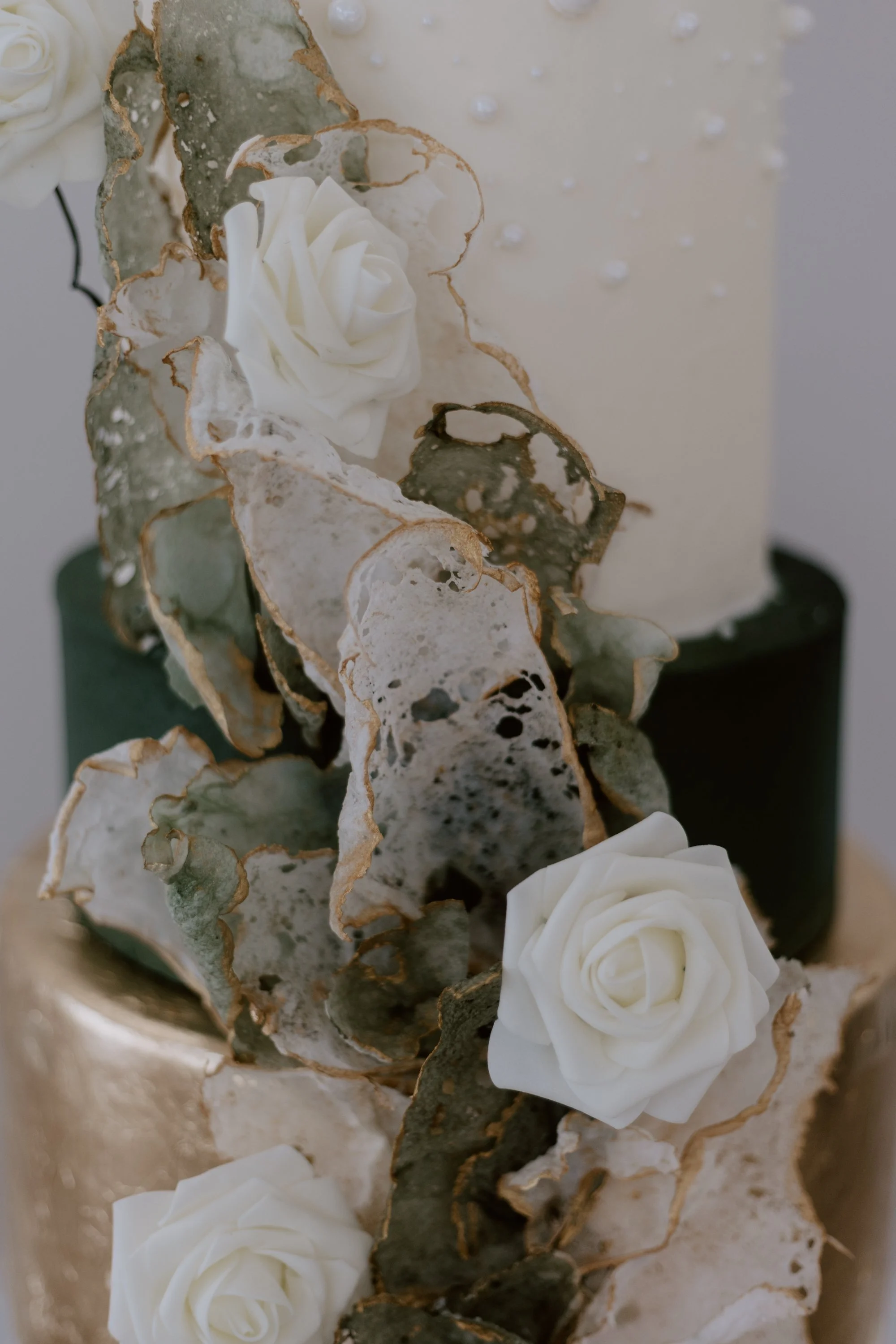 Close-up of a wedding cake decorated with white roses, edible gold leaf, and lace-like sugar details.
