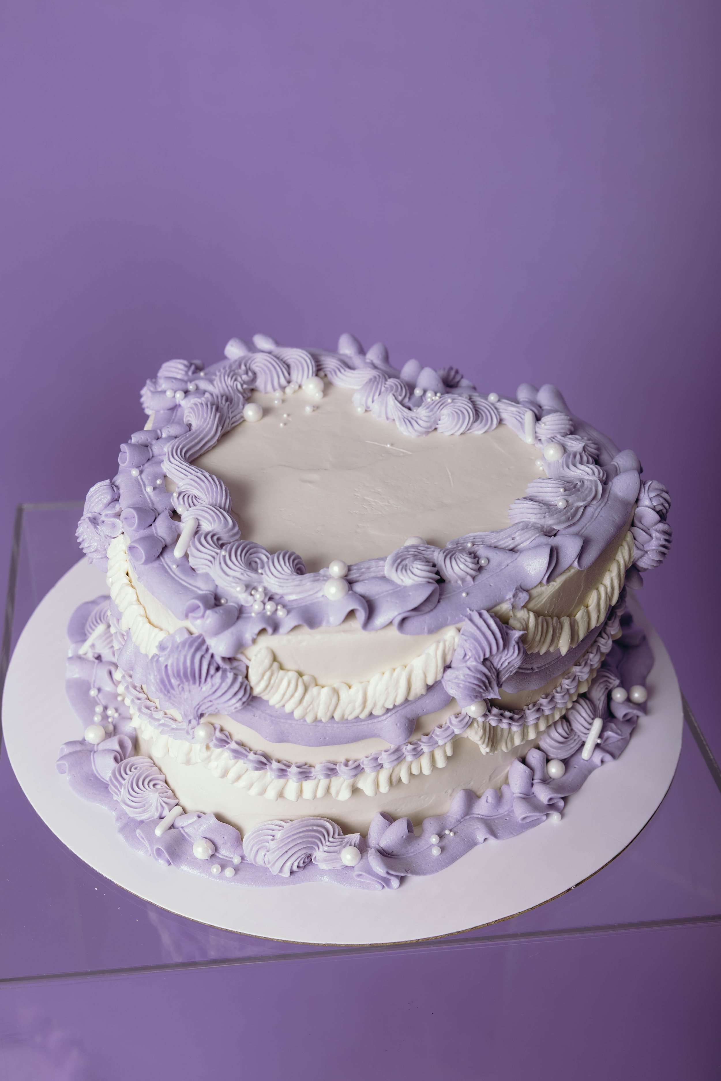 A round cake decorated with purple and white frosting, piped in swirls and decorated with white pearl-like edible beads, on a white cake board against a purple background.