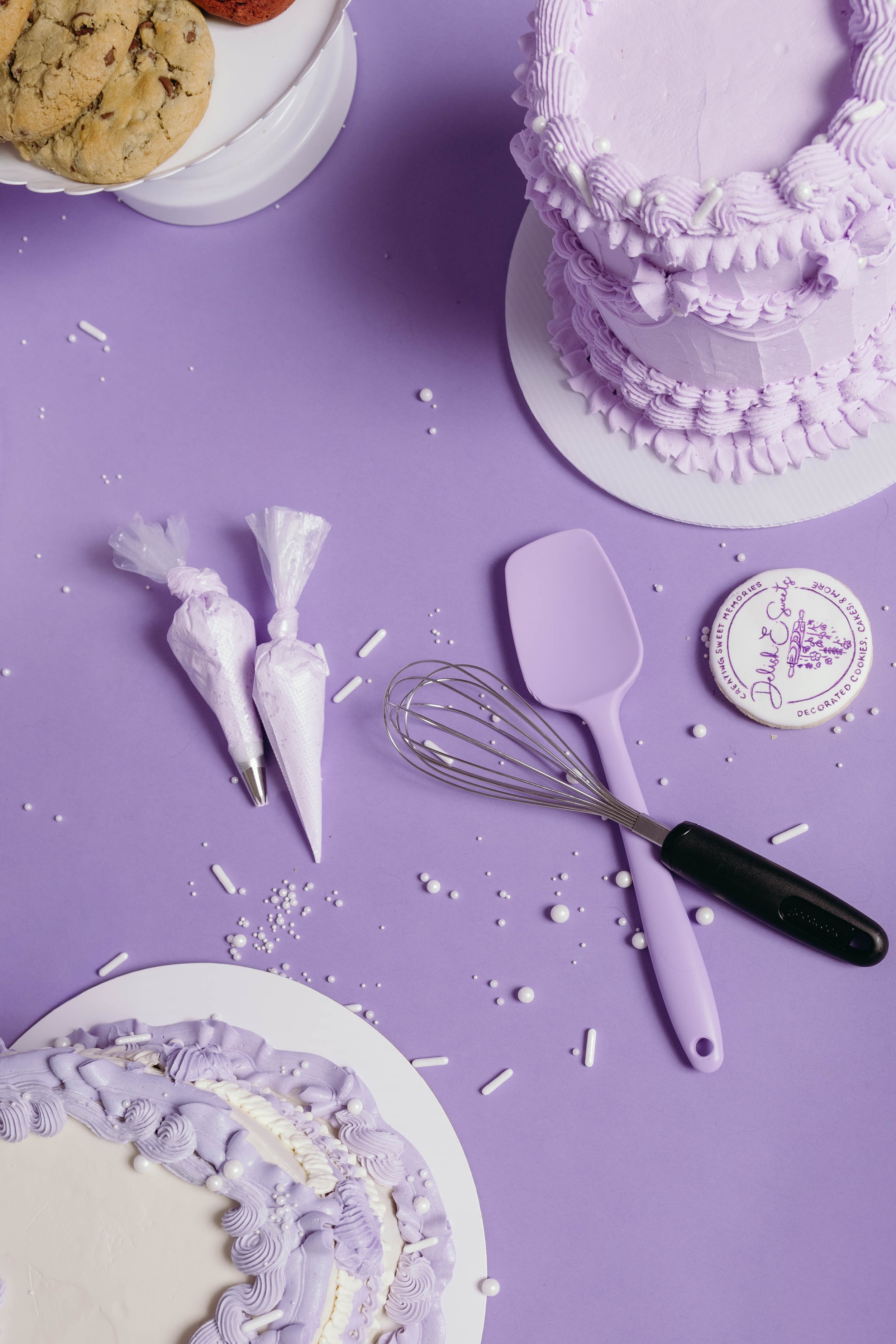 A table decorated with purple and white themed items including a purple frosted cake, cookies, a whisk, and cake decorating tools on a purple background.