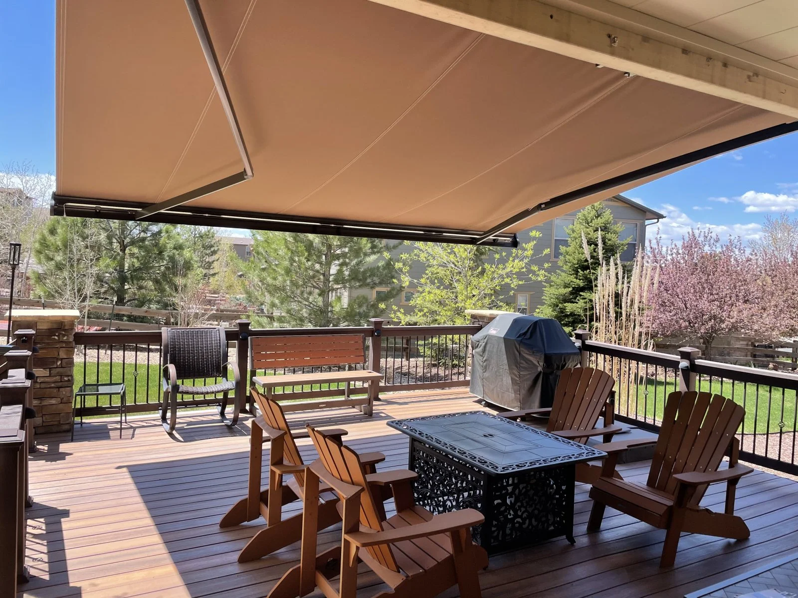 An outdoor deck with wooden flooring, surrounded by trees and greenery under a blue sky. There are chairs, a table, a grill covered with a black cover, and a retractable beige awning overhead.