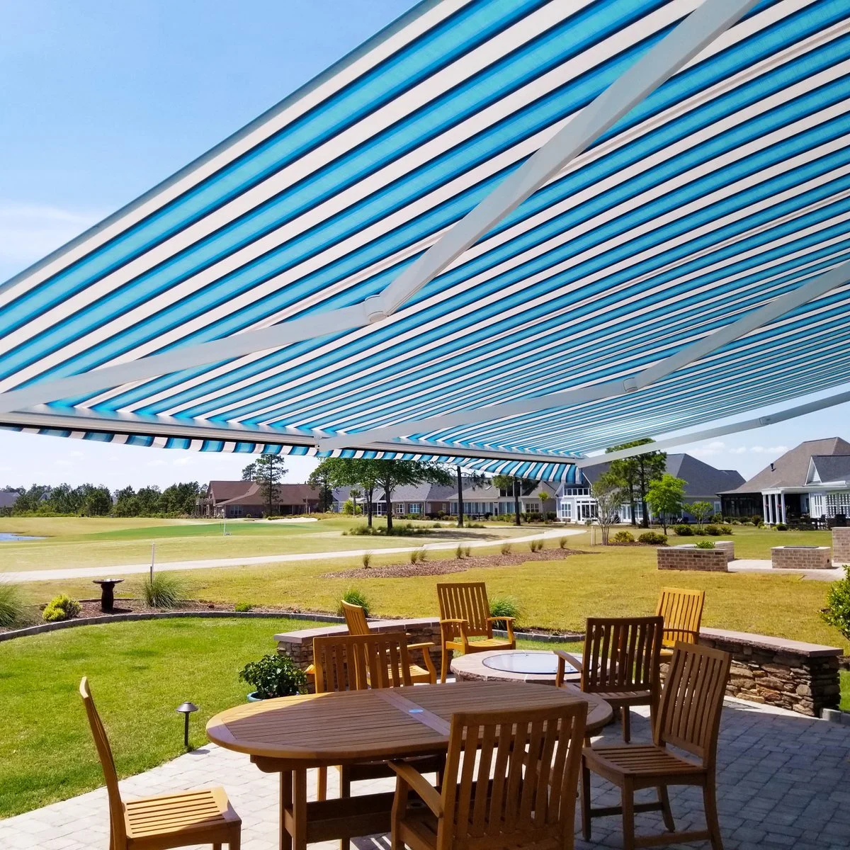 Outdoor patio area with wooden table and chairs under a large blue and white striped retractable awning, overlooking a grassy yard with houses and trees.