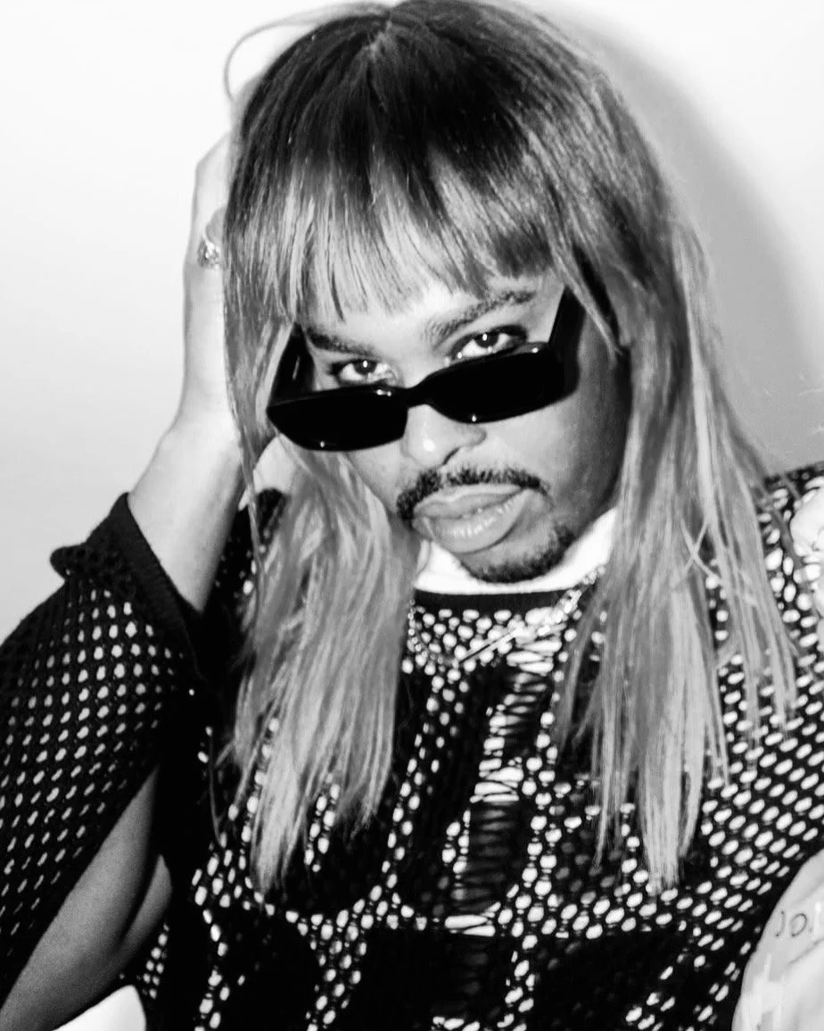 Person with long hair wearing sunglasses and a patterned top, holding their head with one hand, looking at the camera.