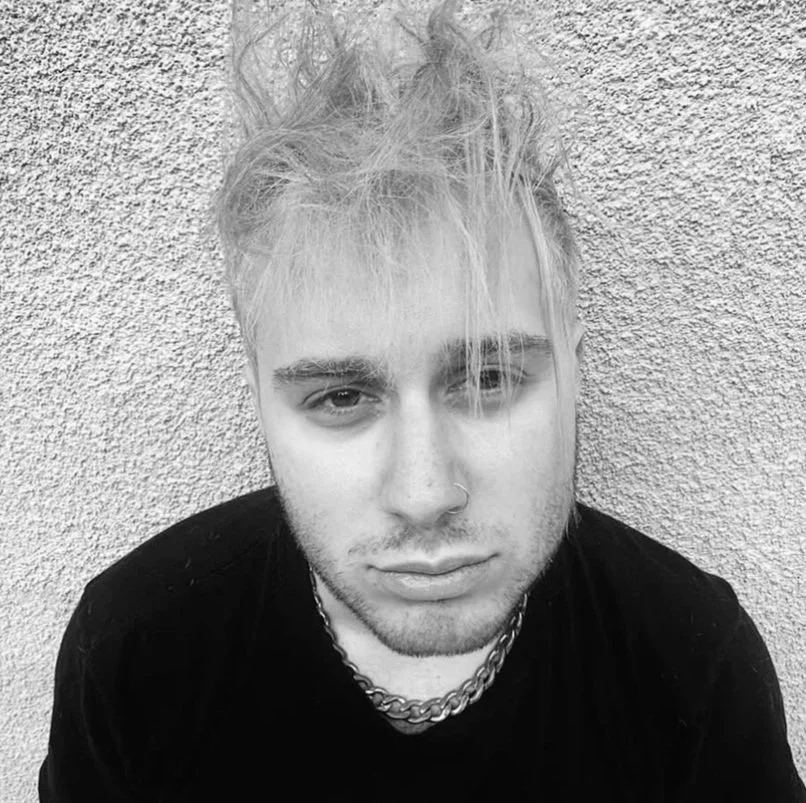 A black and white photo of a young man with messy, light-colored hair, a nose ring, and wearing a chain necklace, against a textured wall background.