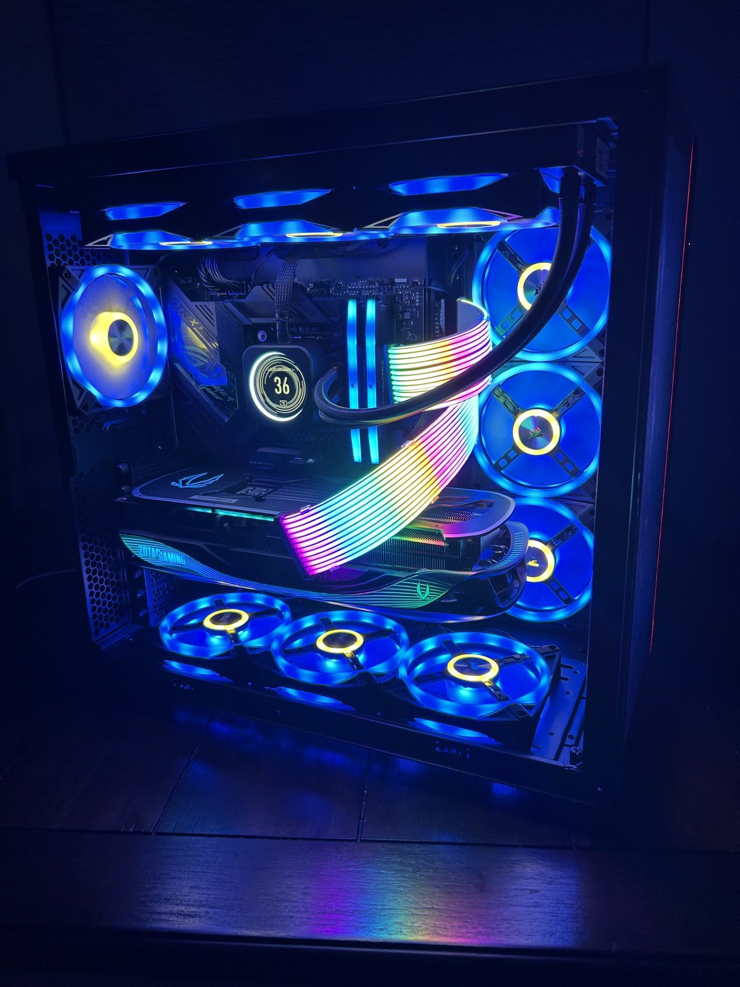 A gaming computer with rainbow RGB lighting, featuring multiple cooling fans, a graphics card, and an illuminated motherboard inside a black case with a transparent side panel.