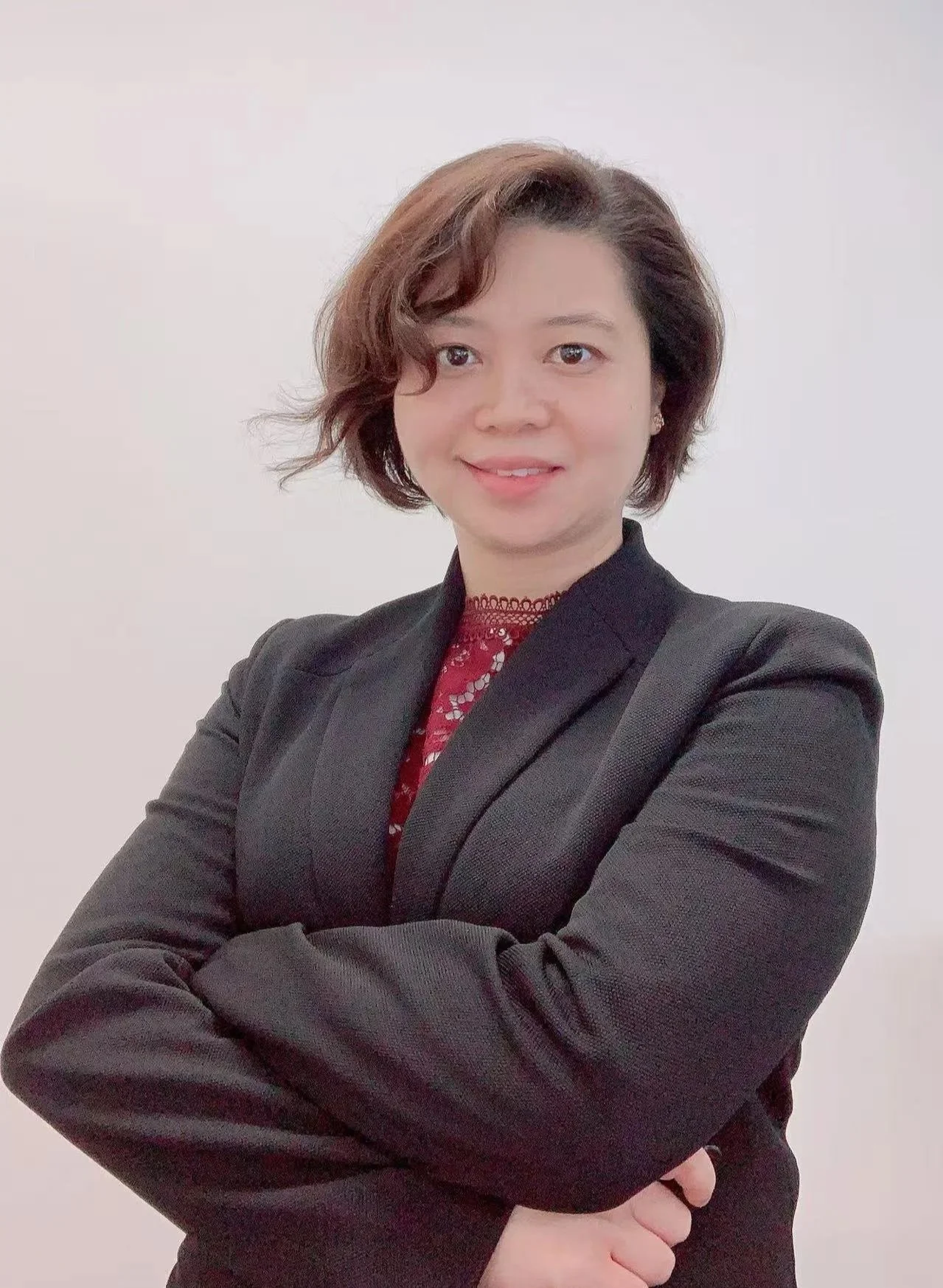 Portrait of a woman with short wavy hair wearing a black blazer over a red lace top, standing against a plain white background, smiling with arms crossed.
