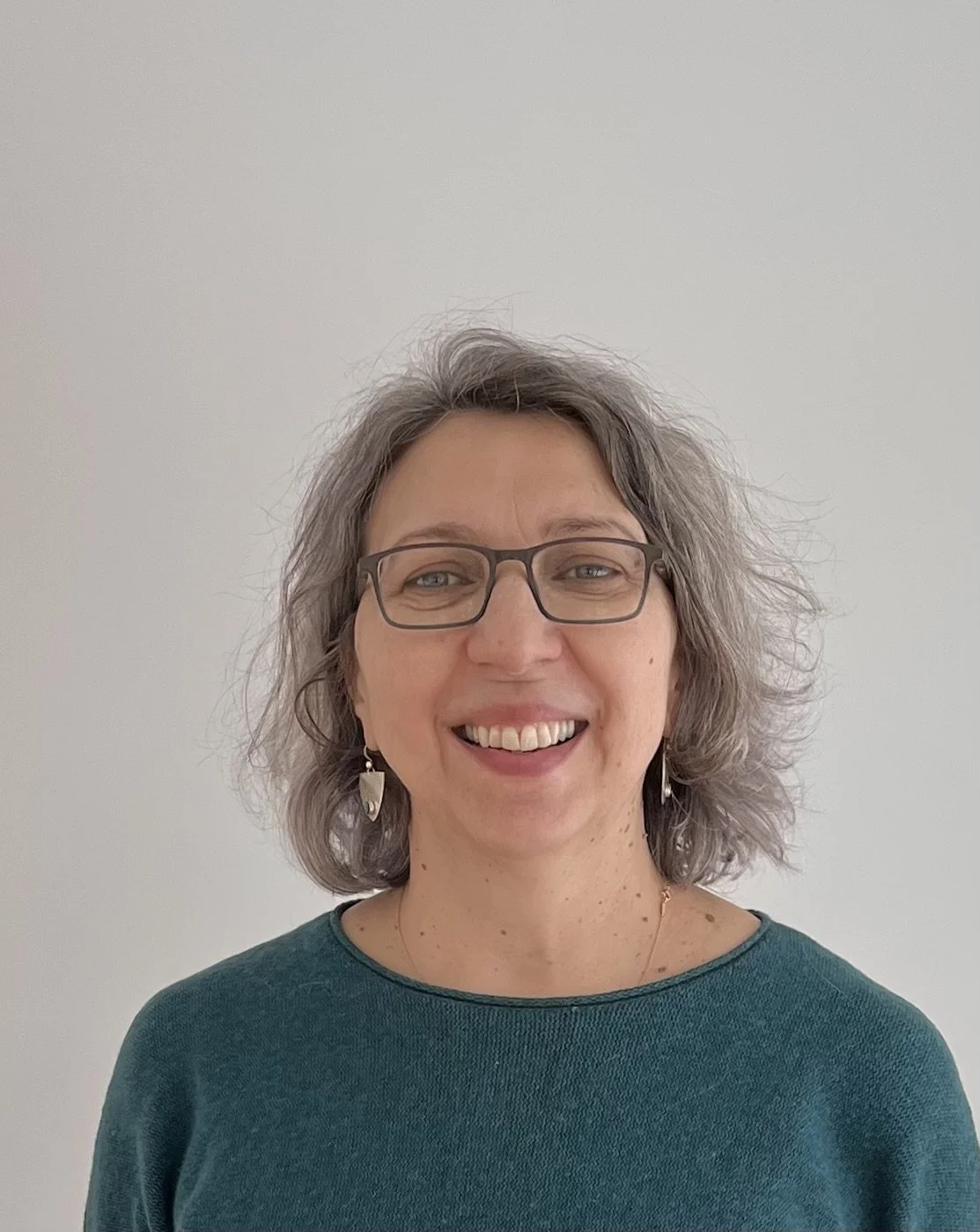A woman with gray hair, wearing glasses, earrings, and a teal top, smiling at the camera against a plain white wall.