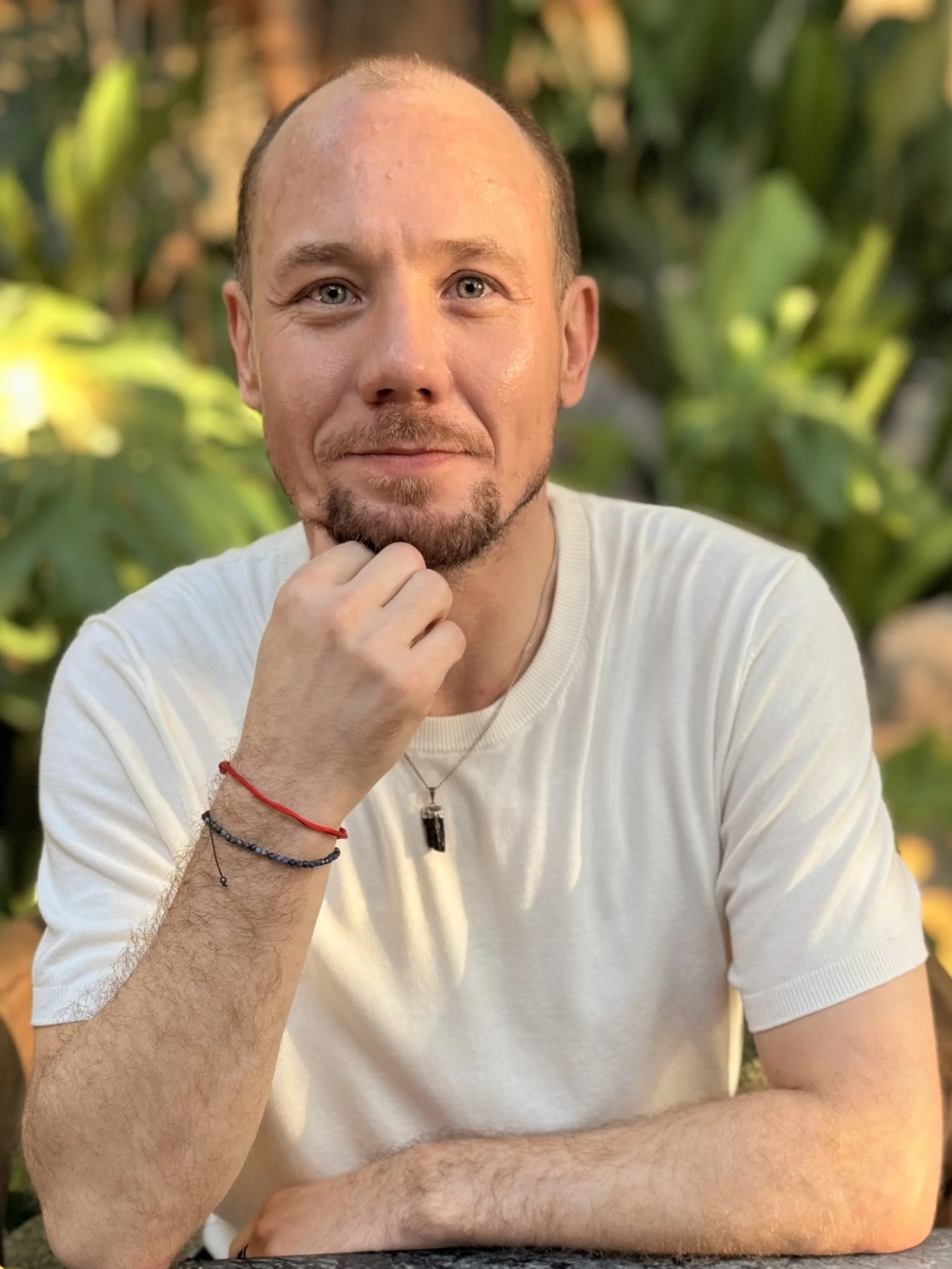 A young man with light skin, short hair, and a goatee, wearing a white shirt, sitting outdoors with a blurred natural background, resting his chin on his hand with bracelets on his wrist.