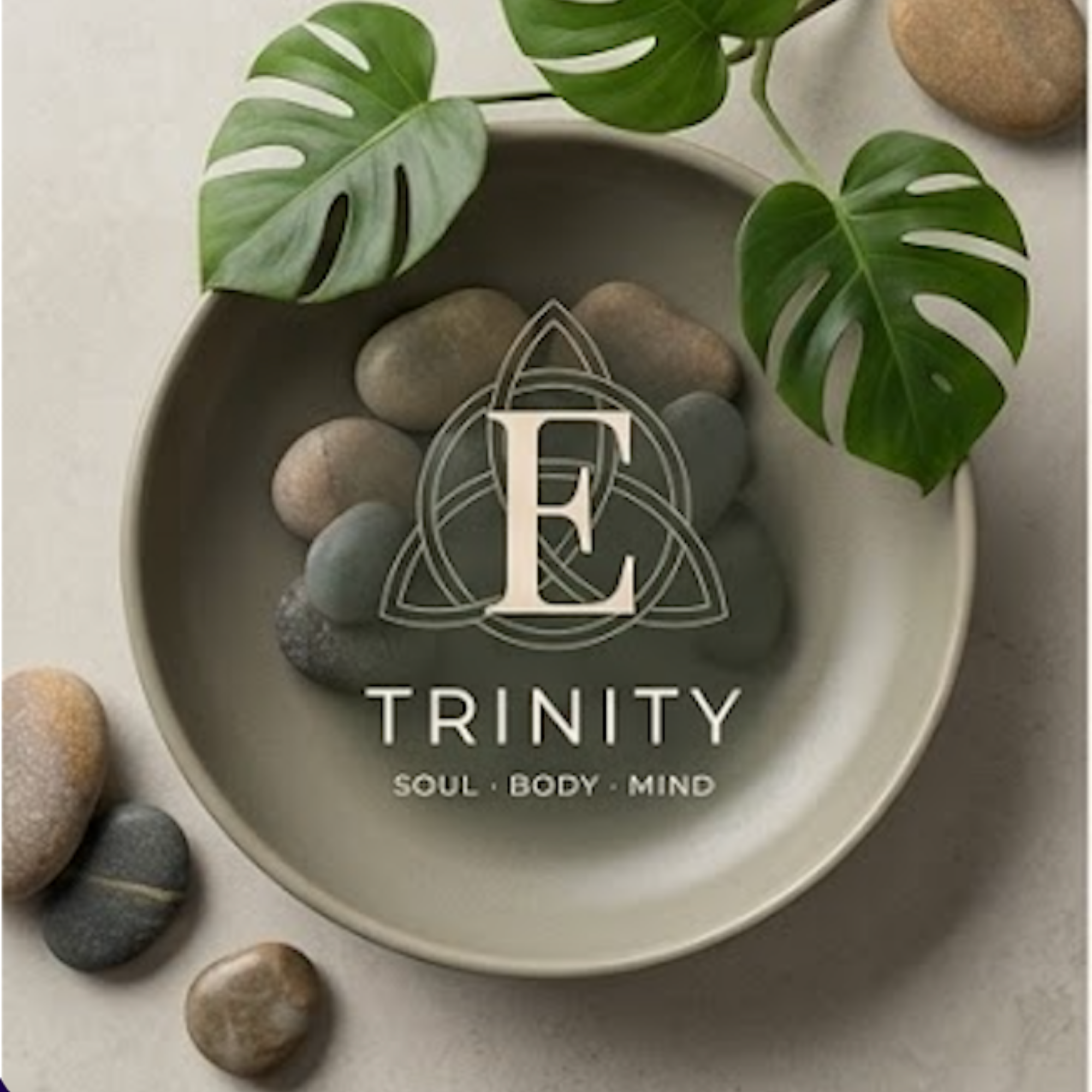 A round gray tray with several smooth, multicolored stones and a white ceramic container with green Monstera leaves peeking over the edge. The tray has a logo with the words 'TRINITY SOUL BODY MIND' and an elegant design around the letter 'T'.
