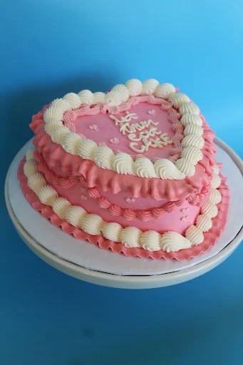 Heart-shaped pink and white birthday cake with 'Happy Valentine' written on top, decorated with ruffled icing and a heart-shaped border.