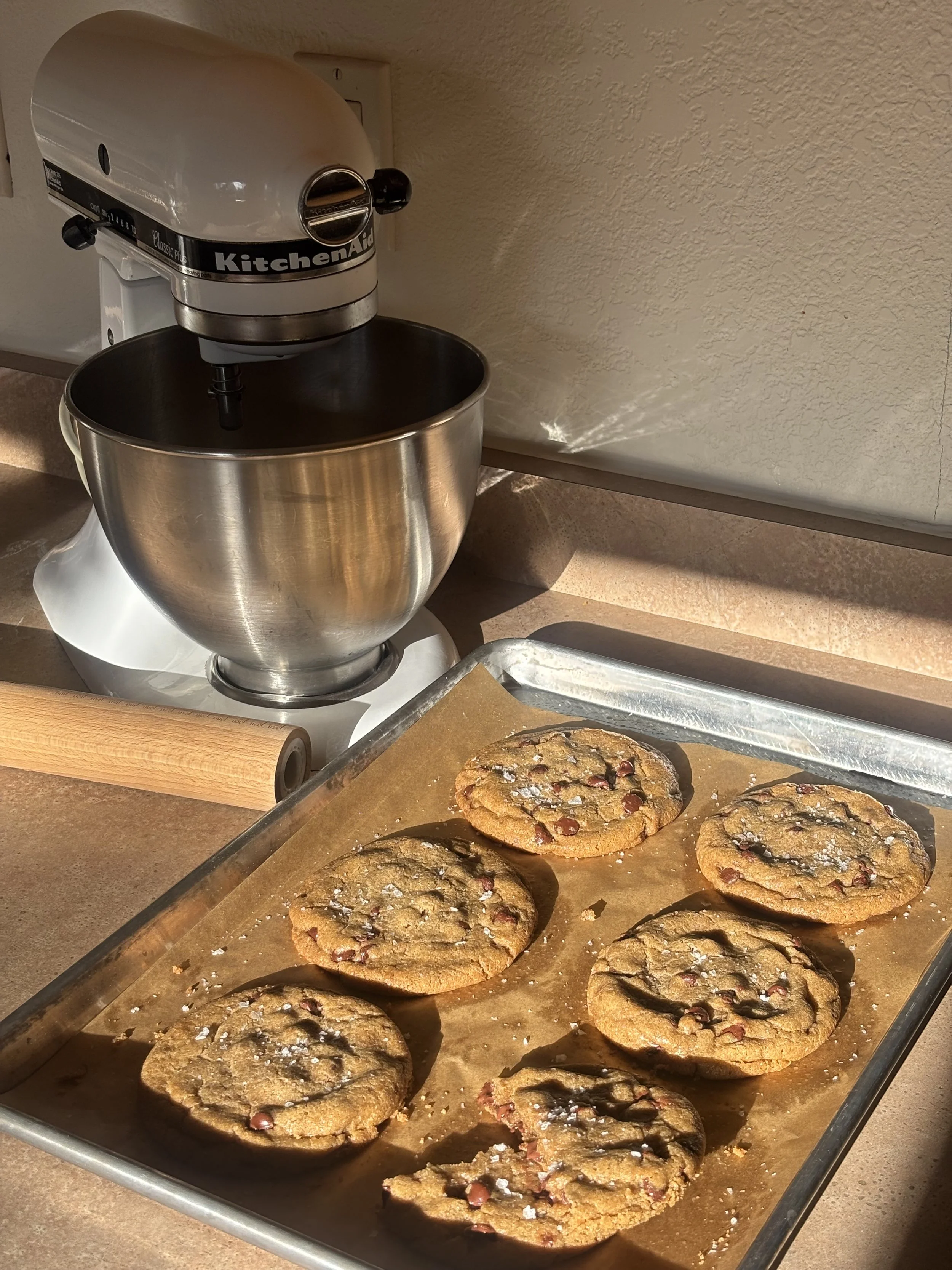 Baked chocolate chip cookies with sea salt on a baking sheet in a kitchen, with a KitchenAid stand mixer in the background.