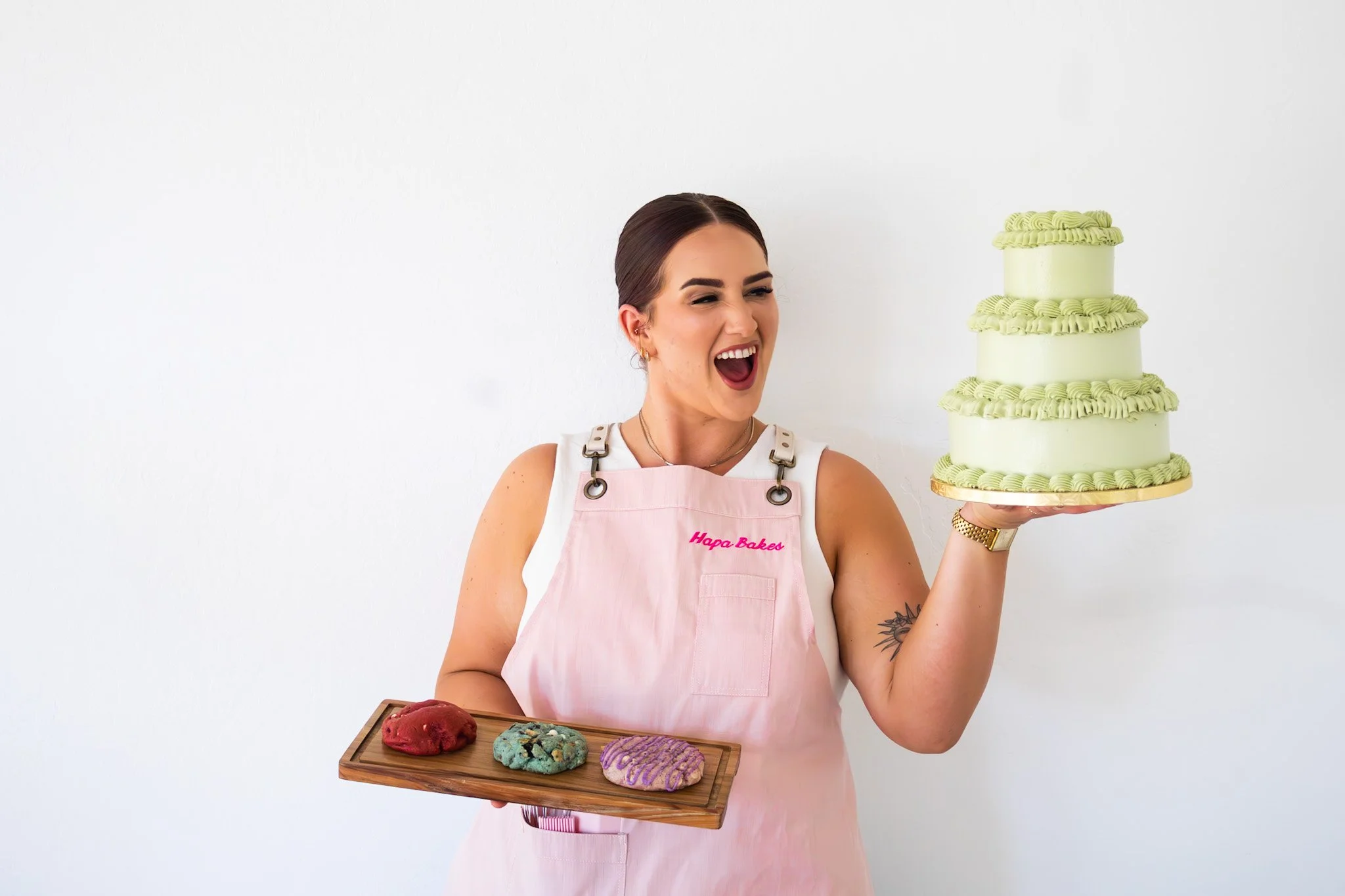 A woman with dark hair and earrings, wearing a pink apron that says "Happie Baked," holding a wooden tray with three colorful cookies and smiling while standing next to a large, layered green cake.
