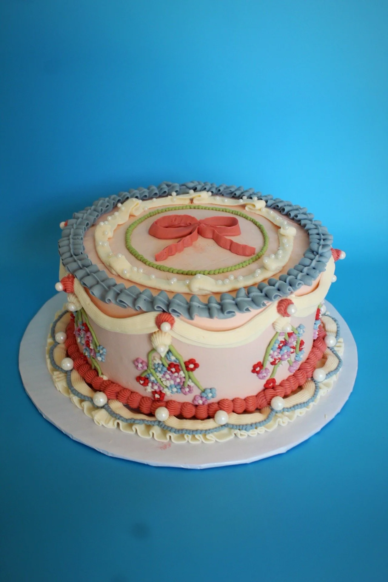 Decorated cake with pink, cream, green, and gray icing, floral designs, pearl accents, and a pink ribbon on top, placed on a white cake board against a blue background.