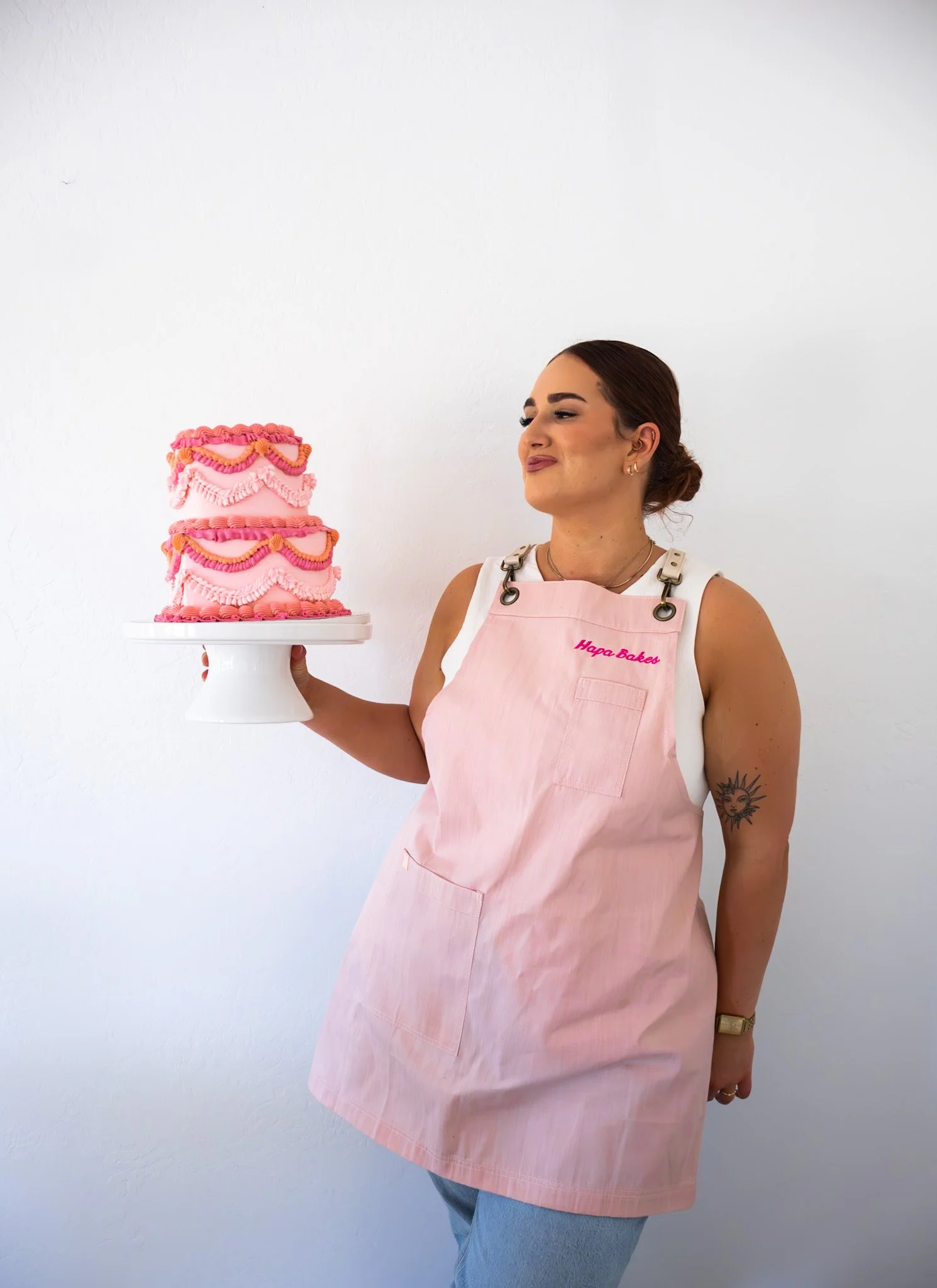 A woman wearing a pink apron holding a decorated pink cake with ruffles, standing against a plain white wall.
