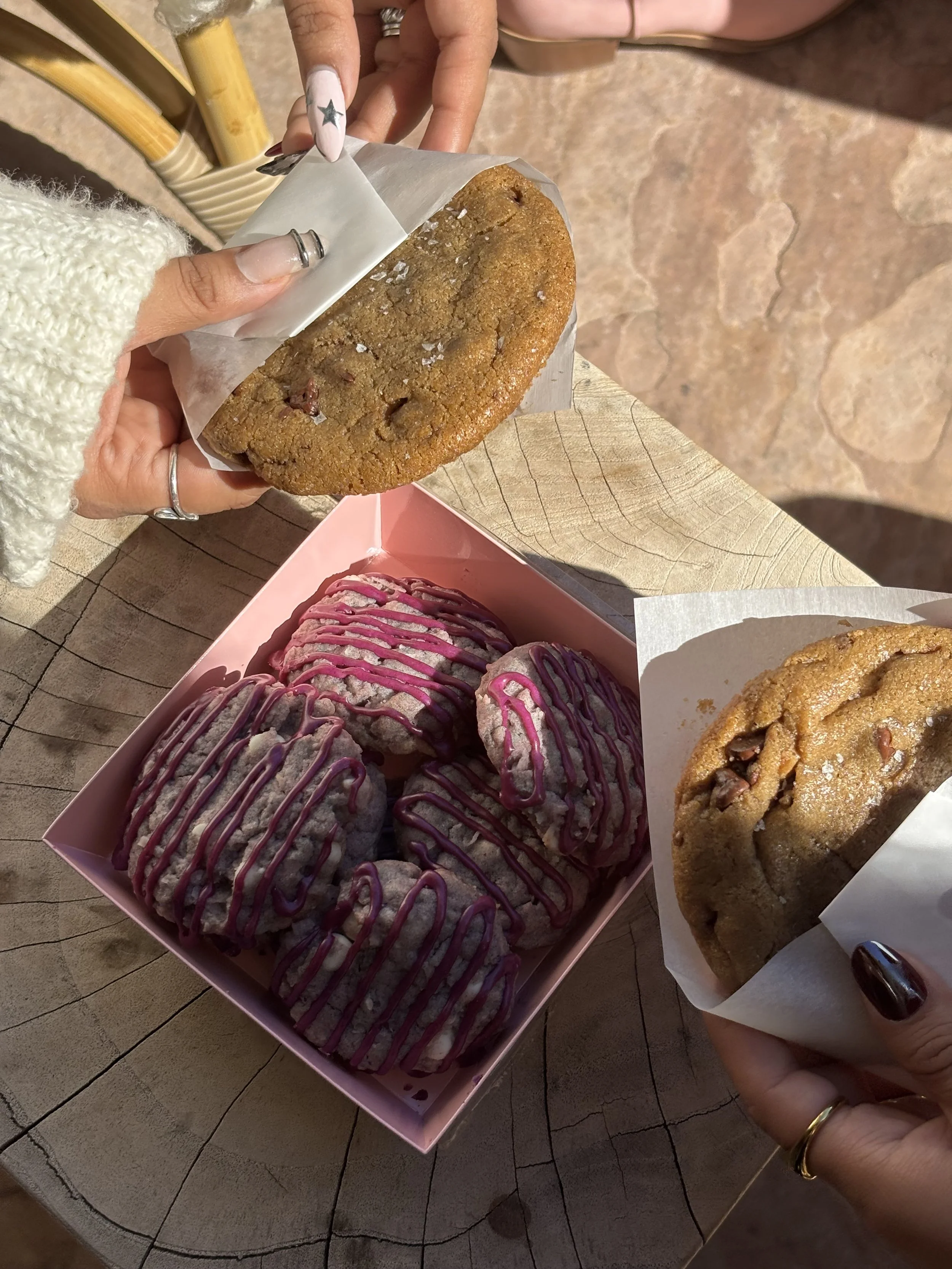 A hand holding a large chocolate chip cookie wrapped in parchment paper, with a pink box of cookies with pink icing and drizzle underneath, and a cookie with pink icing and drizzle on a white paper at the corner.
