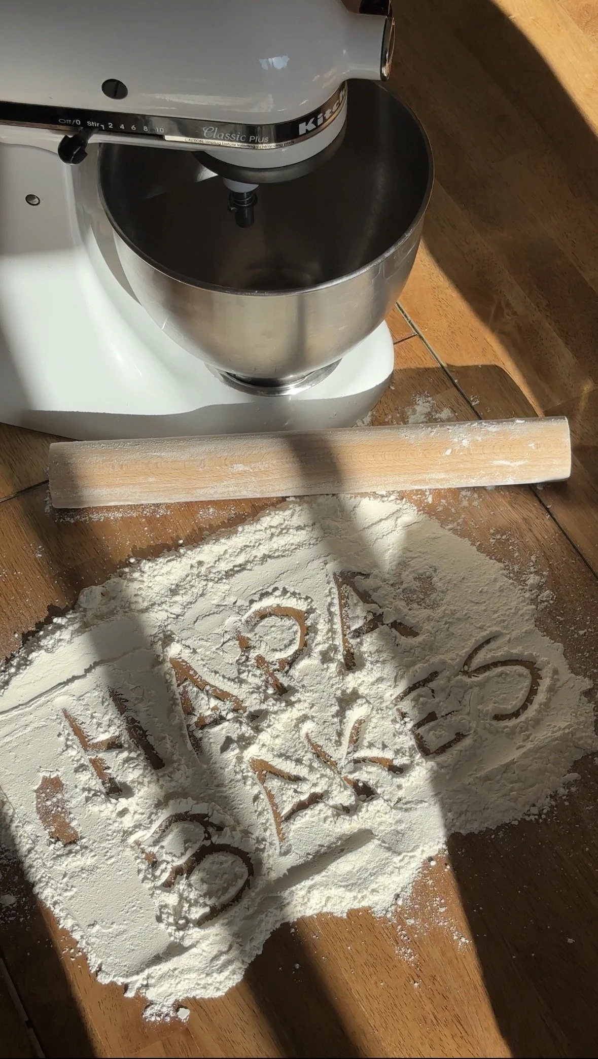 A stand mixer is set on a wooden surface with some flour scattered and a rolling pin nearby. The flour has writing in it, and the surface appears to be used for baking.